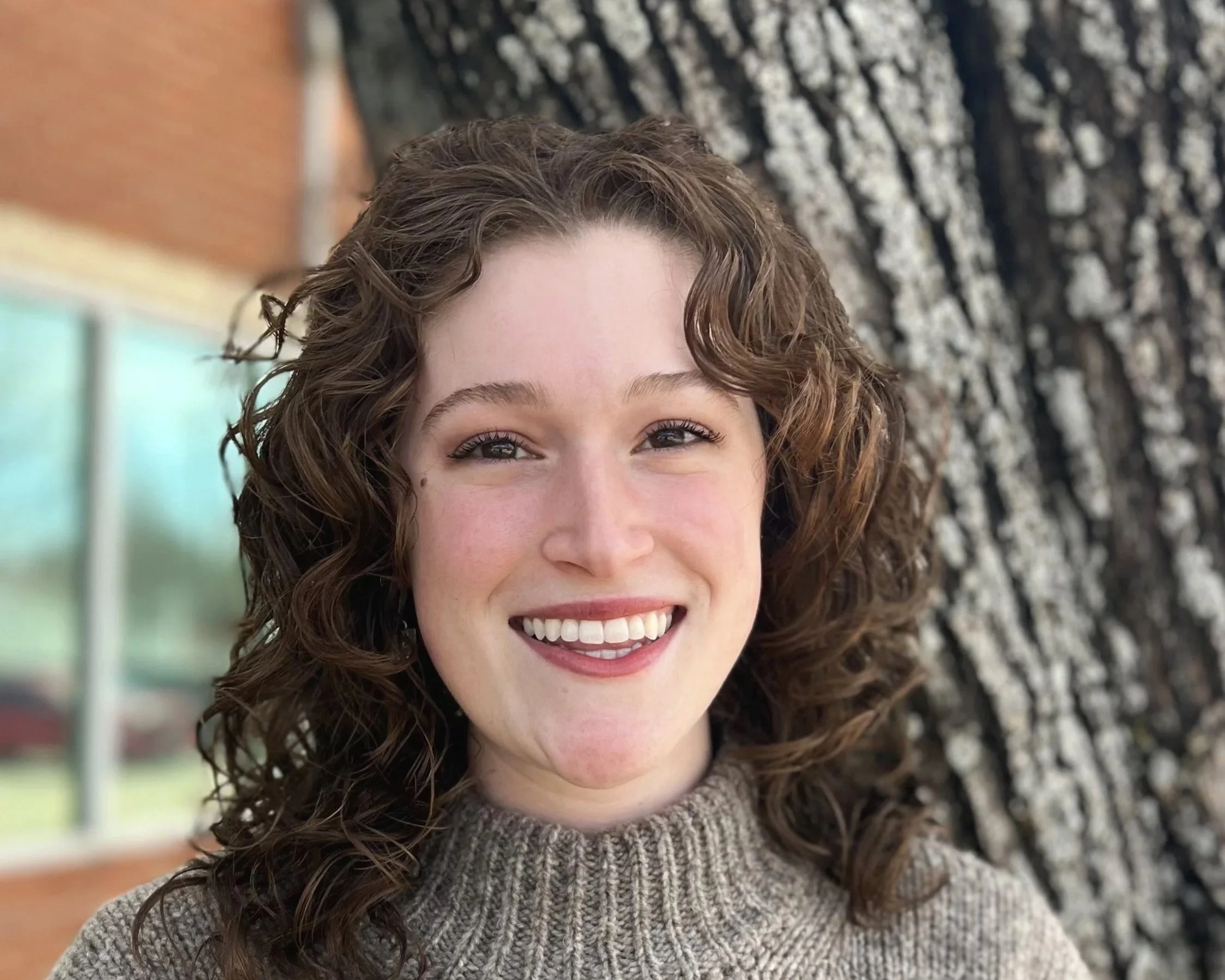 A smiling woman with curly brown hair wearing a beige knit sweater standing outside near a large tree with textured bark.