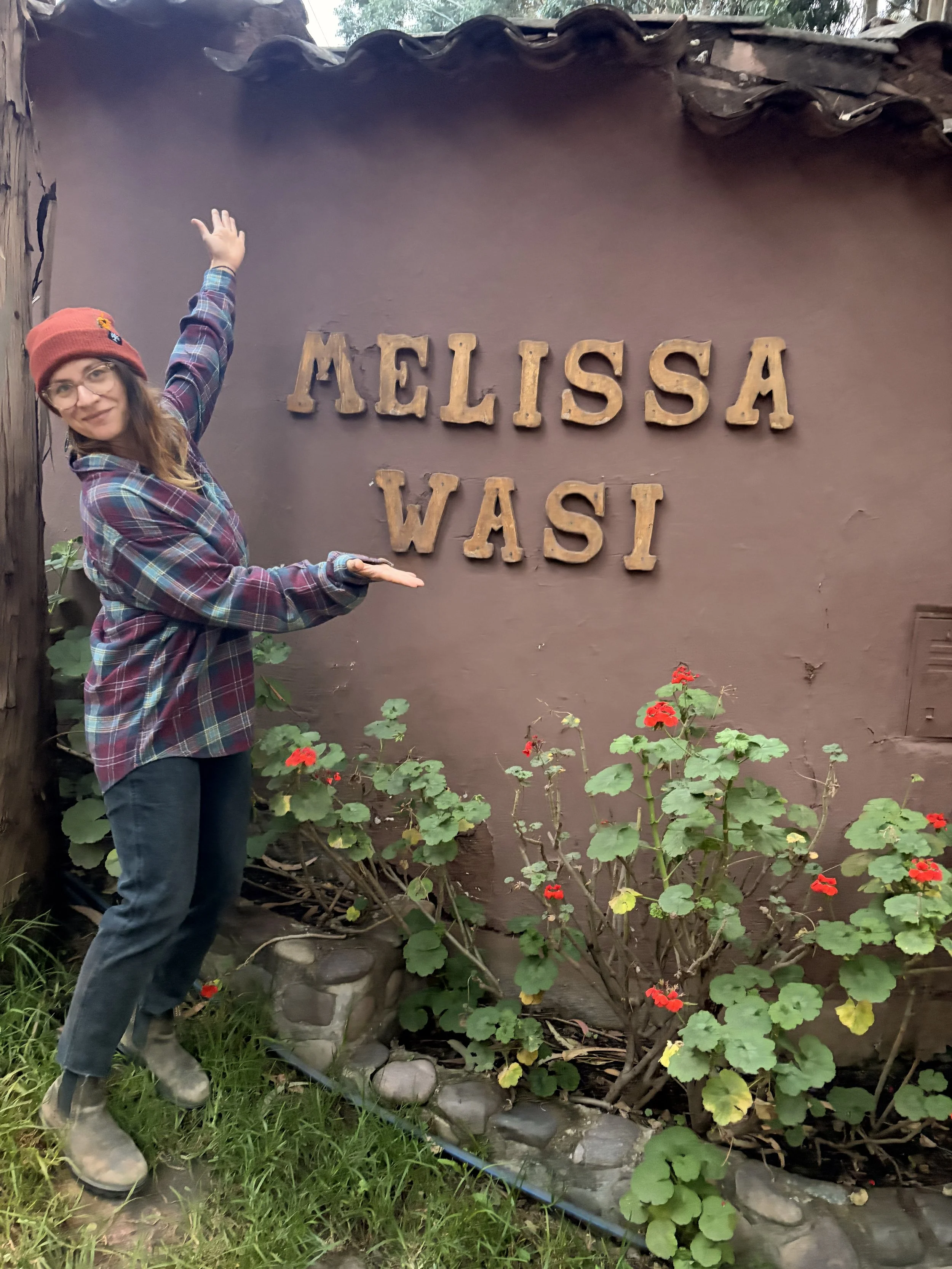 A woman in glasses, a red beanie, and a plaid jacket points to a sign that reads 'Melissa Wasi' on a brown wall. There are red flowers and green leaves in front of the wall, and she is standing outdoors on grass.