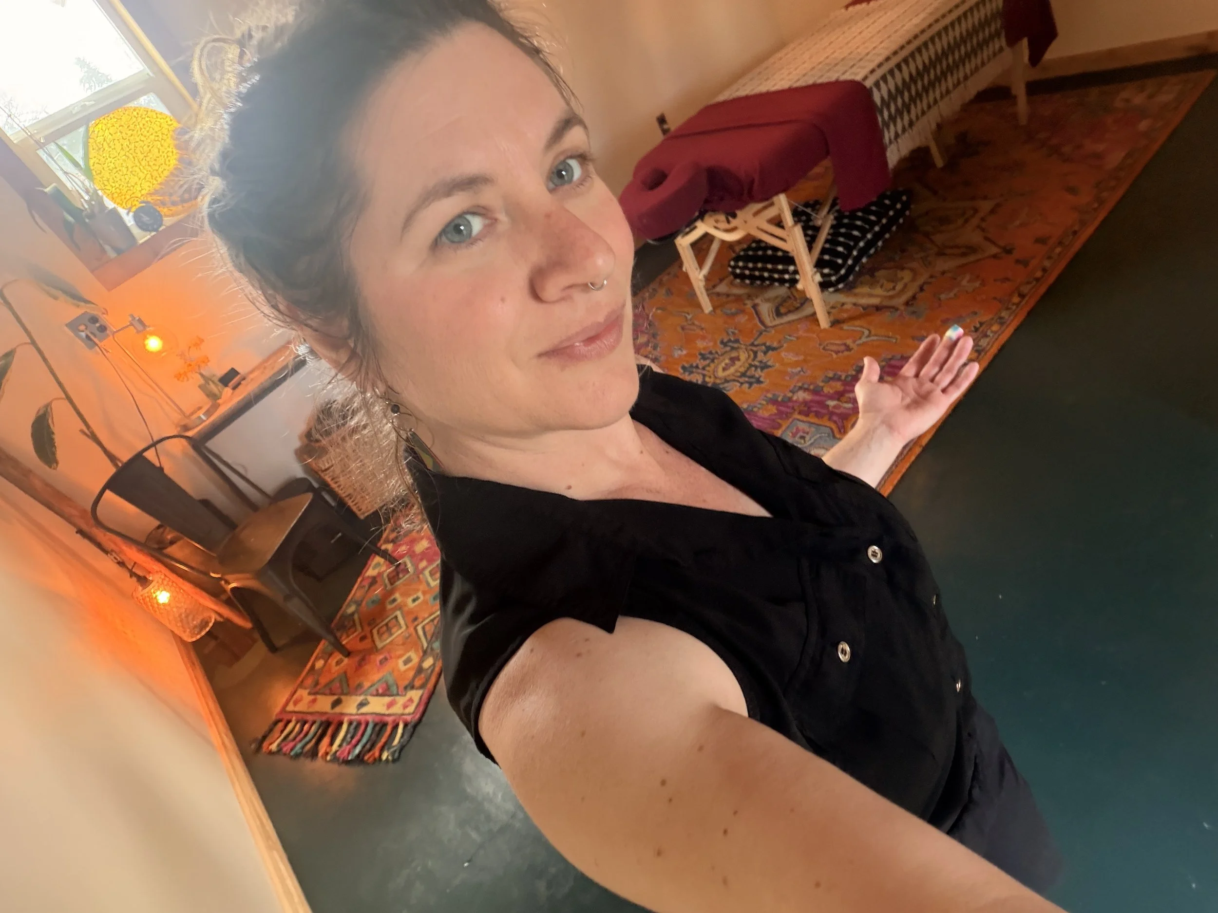 A woman with blue eyes, dark hair in a messy bun, and a septum piercing taking a selfie in a room with colorful decor. In the background, there is a patterned rug, a folding chair, a red blanket, a window with sunlight, and a wall with artwork and hanging plants.