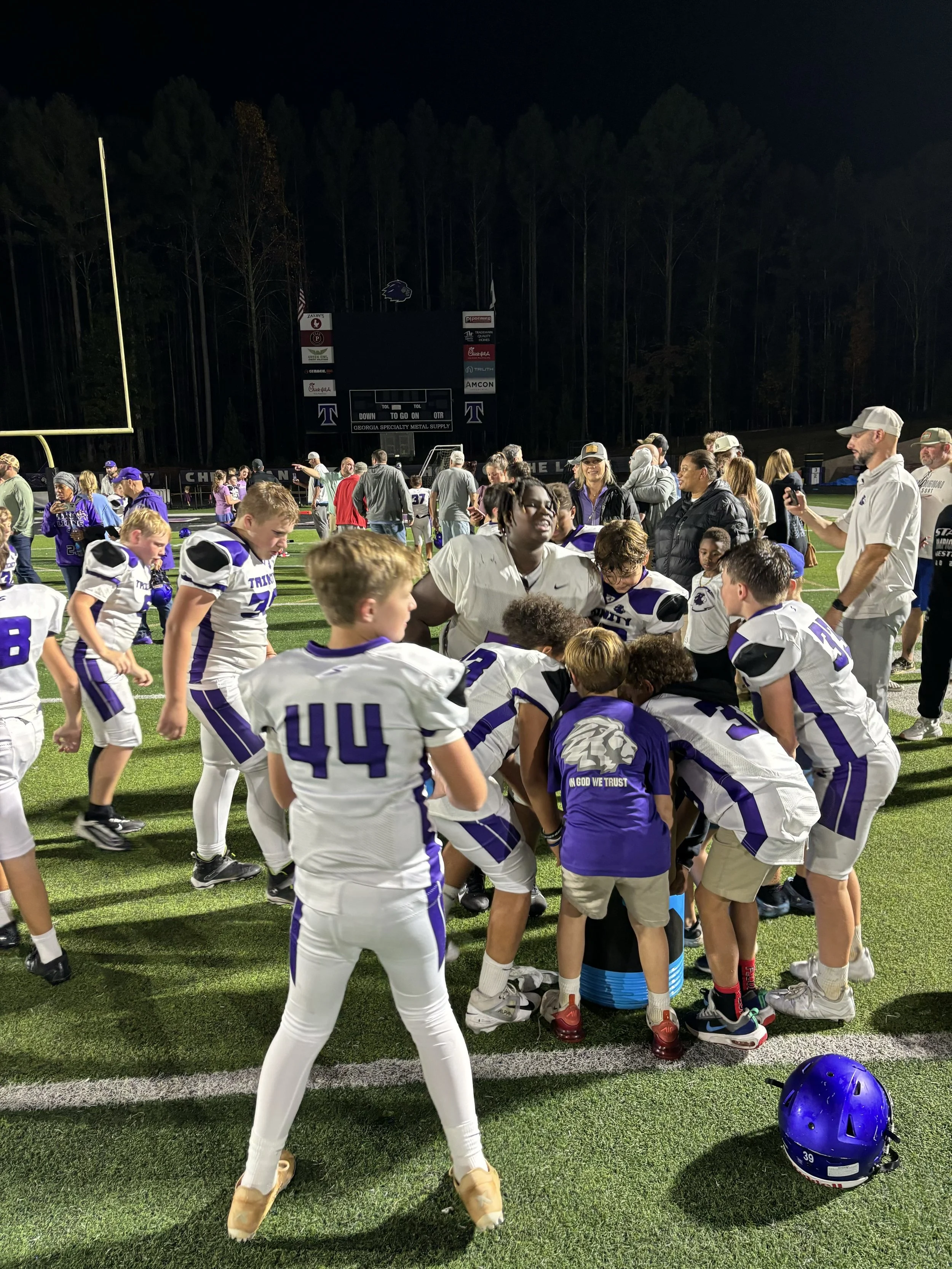 Trinity Christian School 5th/6th grade football players huddle with teammates and a young fan after their Fall 2024 championship win.