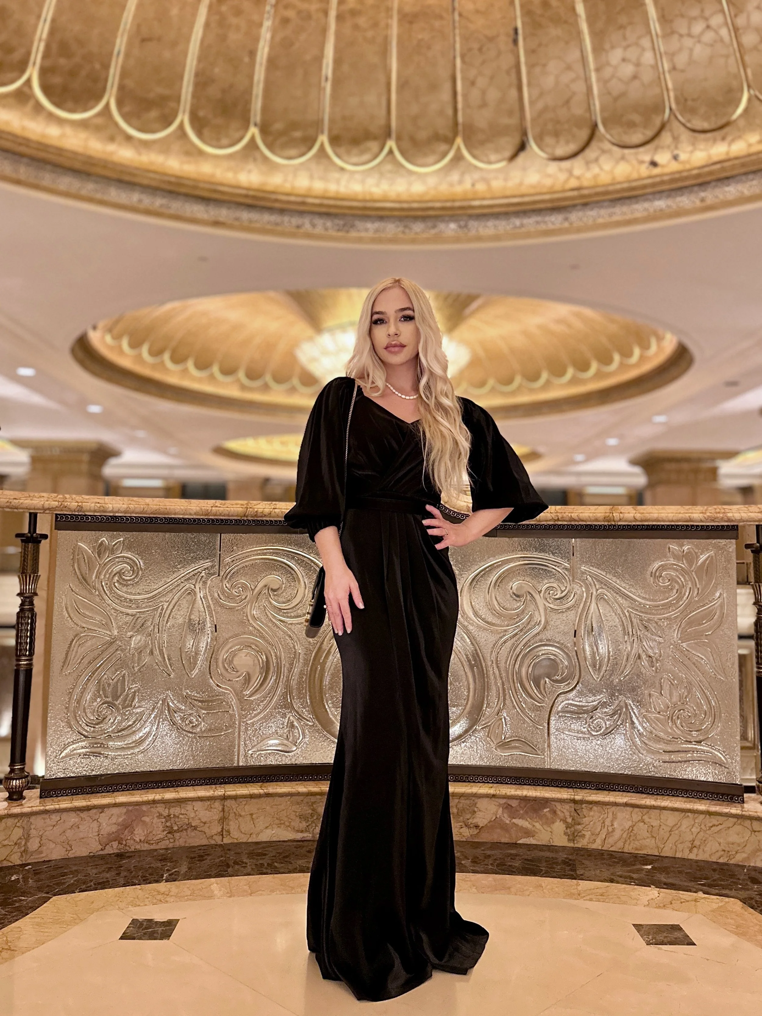 A young woman with long blonde wavy hair wearing a black strapless dress with puffy sleeves, a pearl necklace, and gloves, standing in an ornate, gold-accented lobby with a decorative marble railing and a large, elaborate ceiling chandelier.