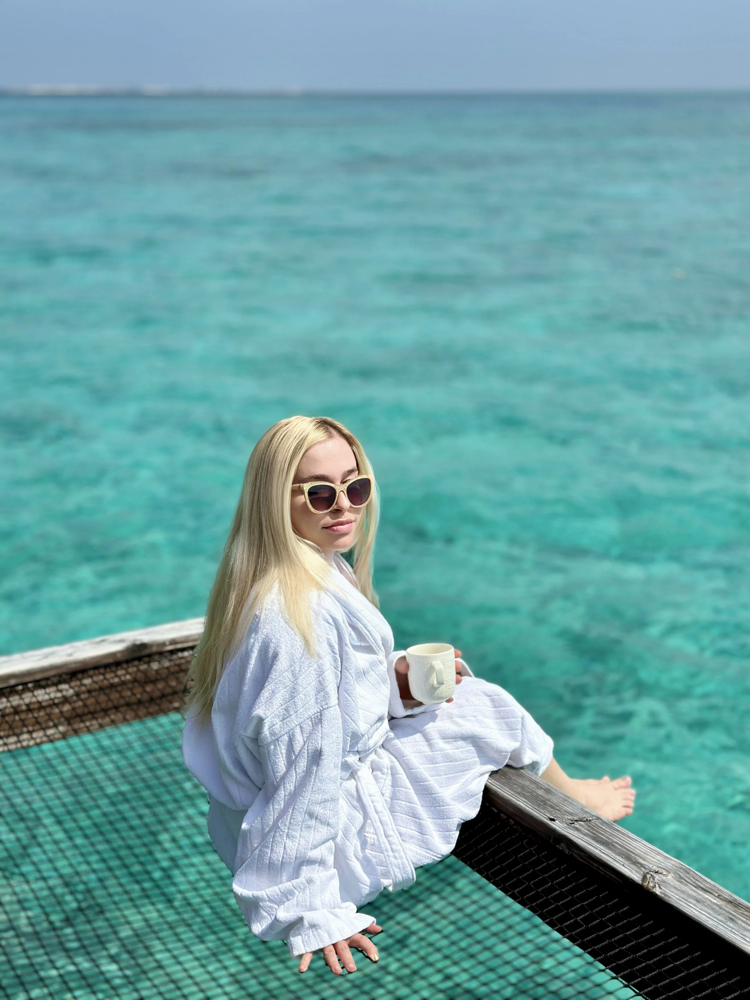 A woman with long blonde hair, wearing large sunglasses and a white robe, sitting on a wooden deck near clear turquoise ocean water, holding a white mug.