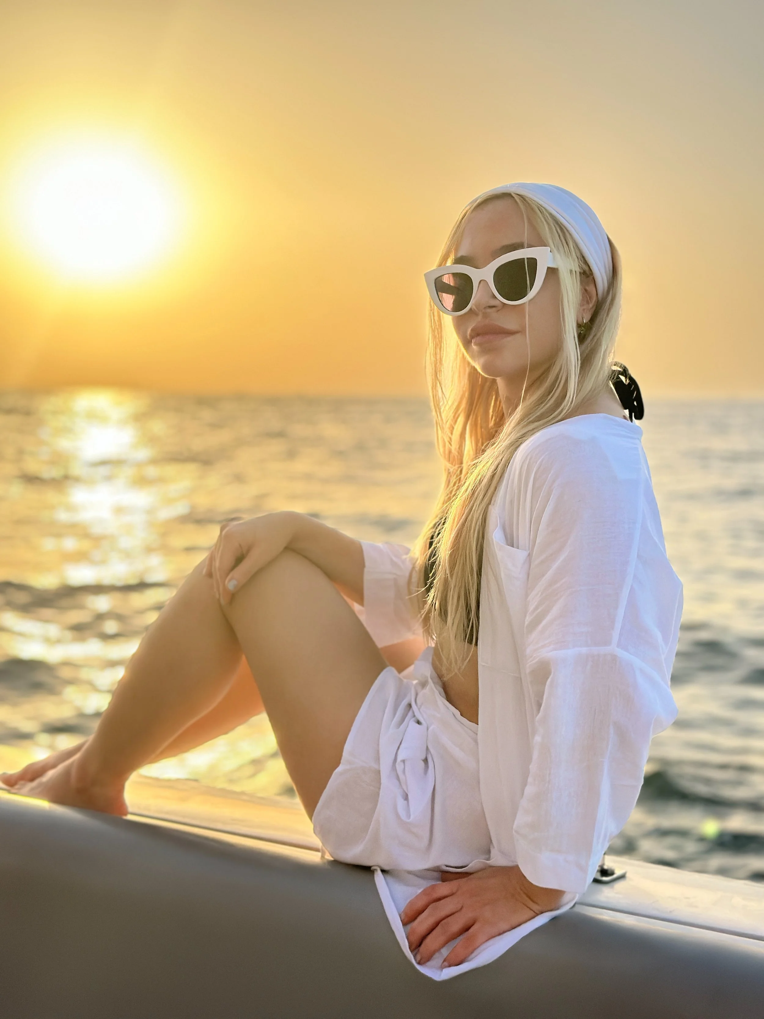 A woman with blonde hair wearing white sunglasses and a white headscarf sitting near water during sunset, with the sun low in the sky.