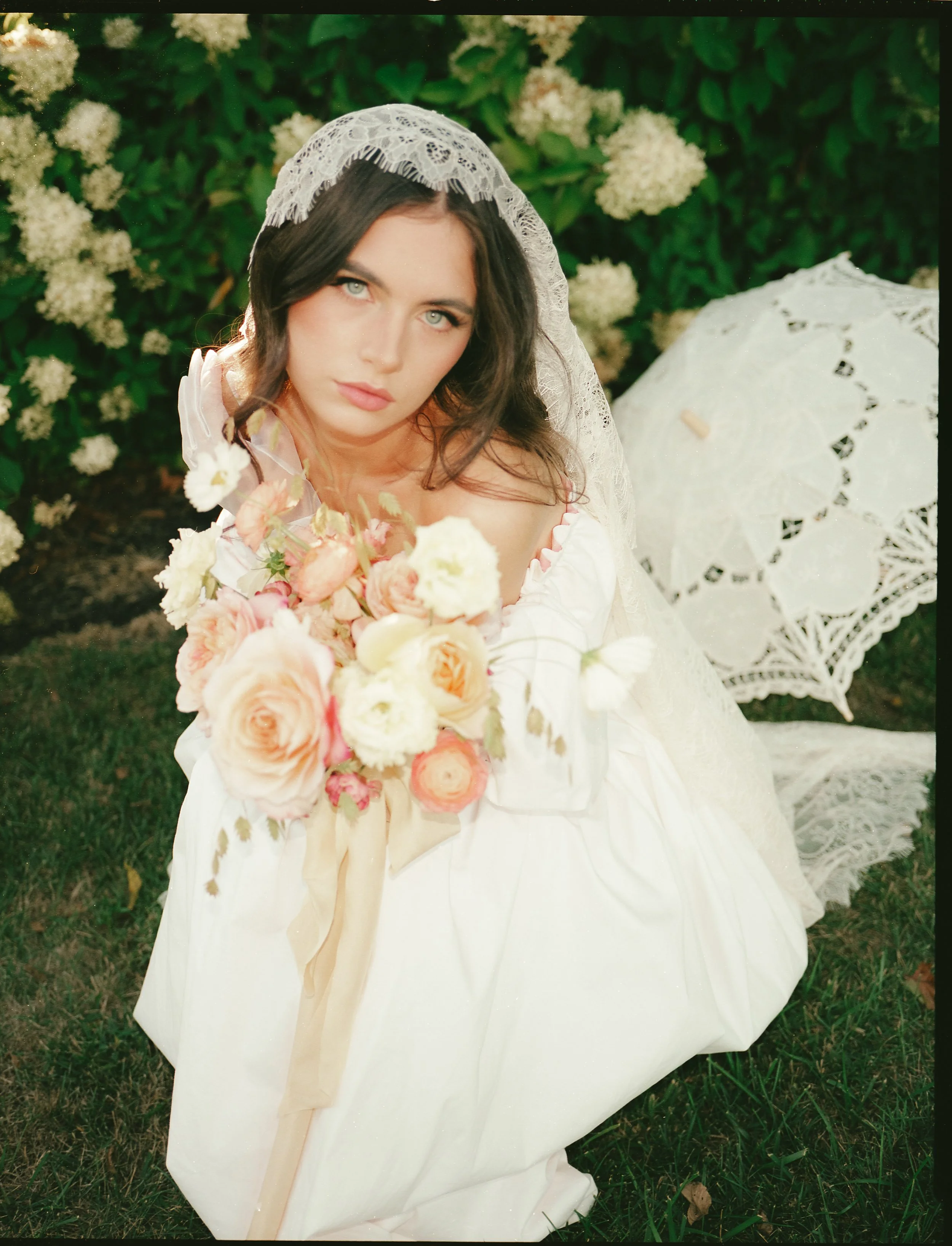 A woman in a vintage wedding dress with an off-the-shoulder neckline, holding a large bouquet of blush, peach, and cream roses. She has shoulder-length dark hair with loose waves, blue eyes, and a lace veil. She sits on the grass in front of a bush with white flowers, with a white lace parasol nearby.
