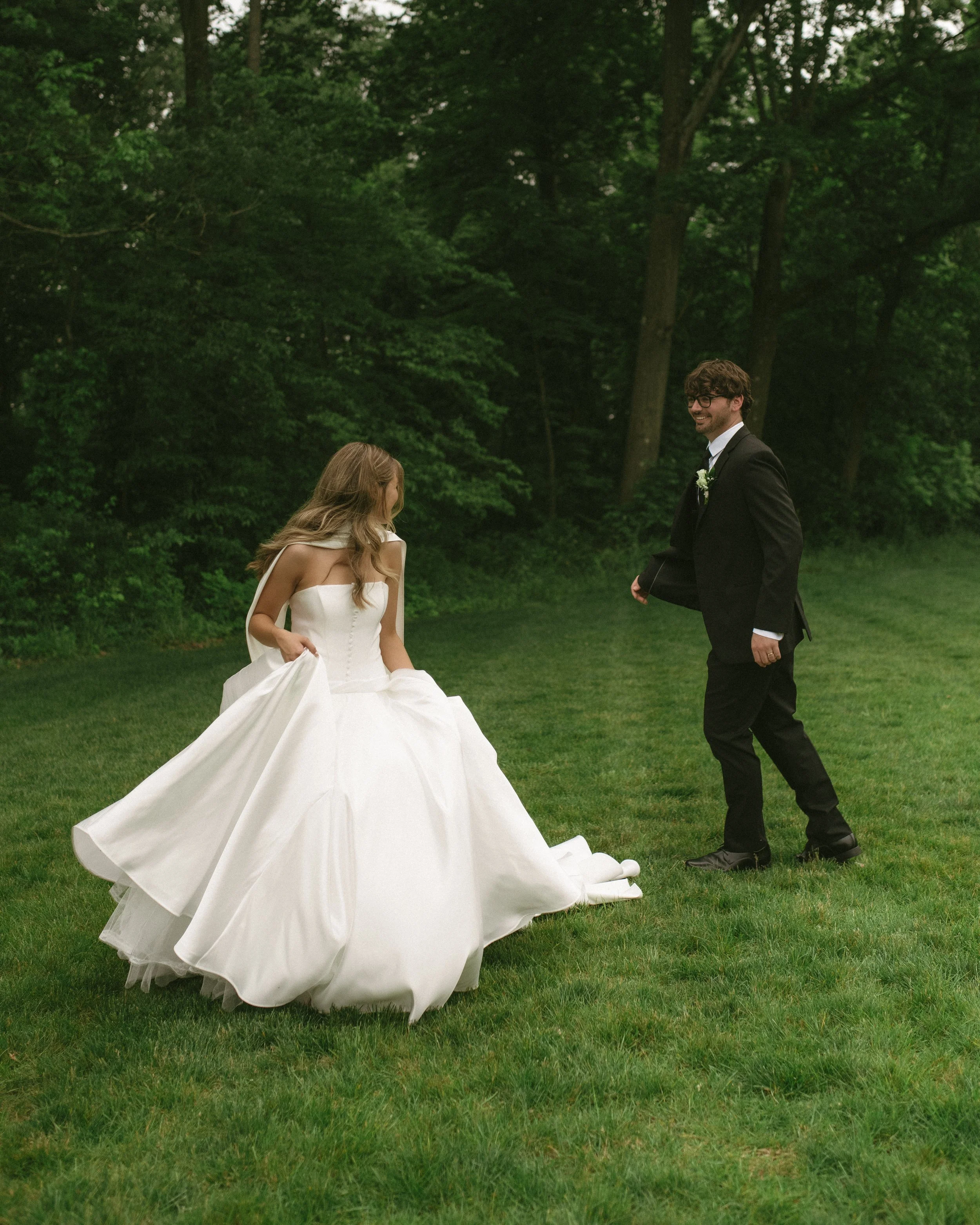 A bride in a white wedding gown and a groom in a black tuxedo stand on a grassy area surrounded by trees, with the bride holding her dress and the groom smiling as they look at each other.