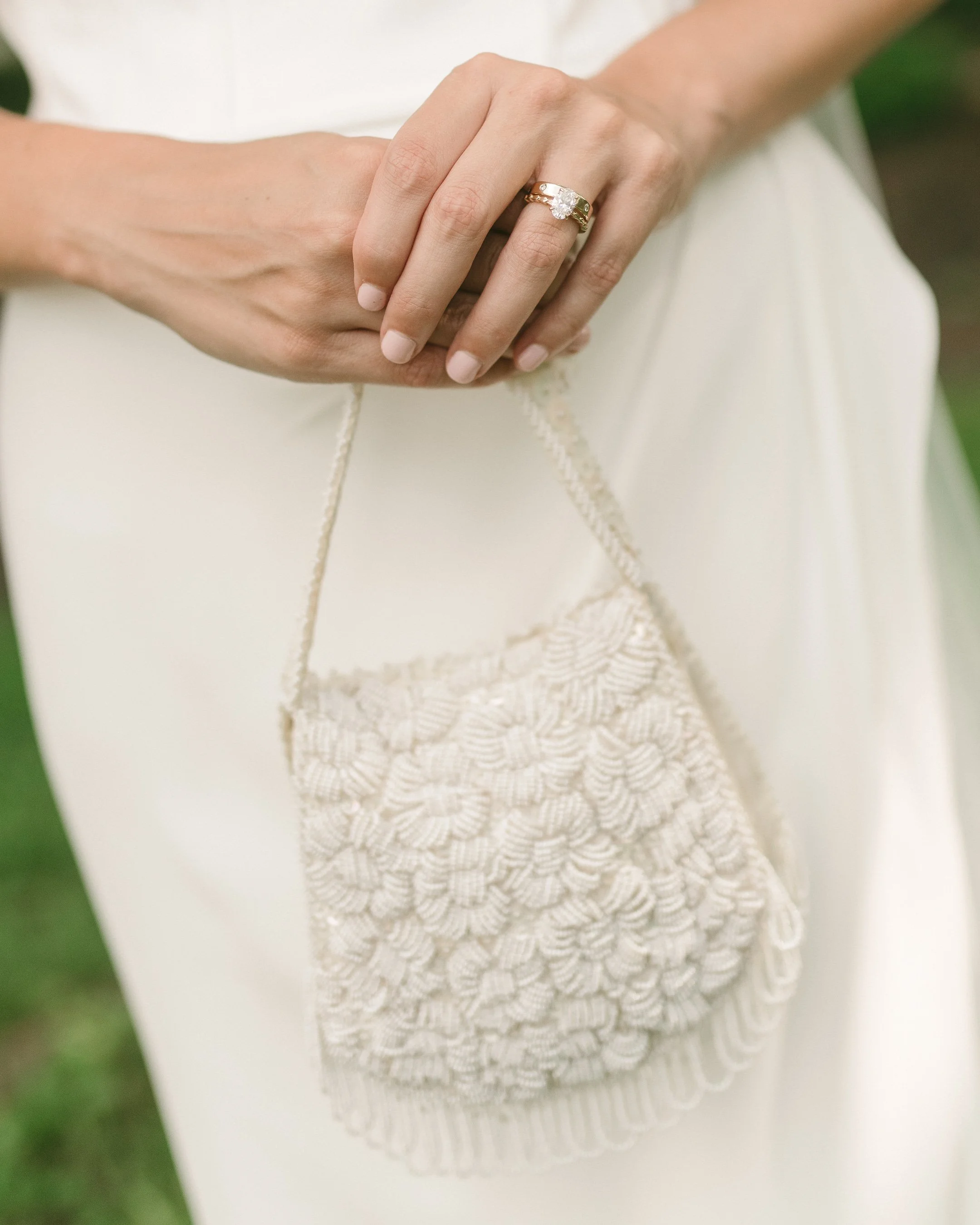 Close-up of a woman's hands holding a small, embroidered cream-colored purse, wearing a gold ring with a large gemstone, and dressed in a white gown.