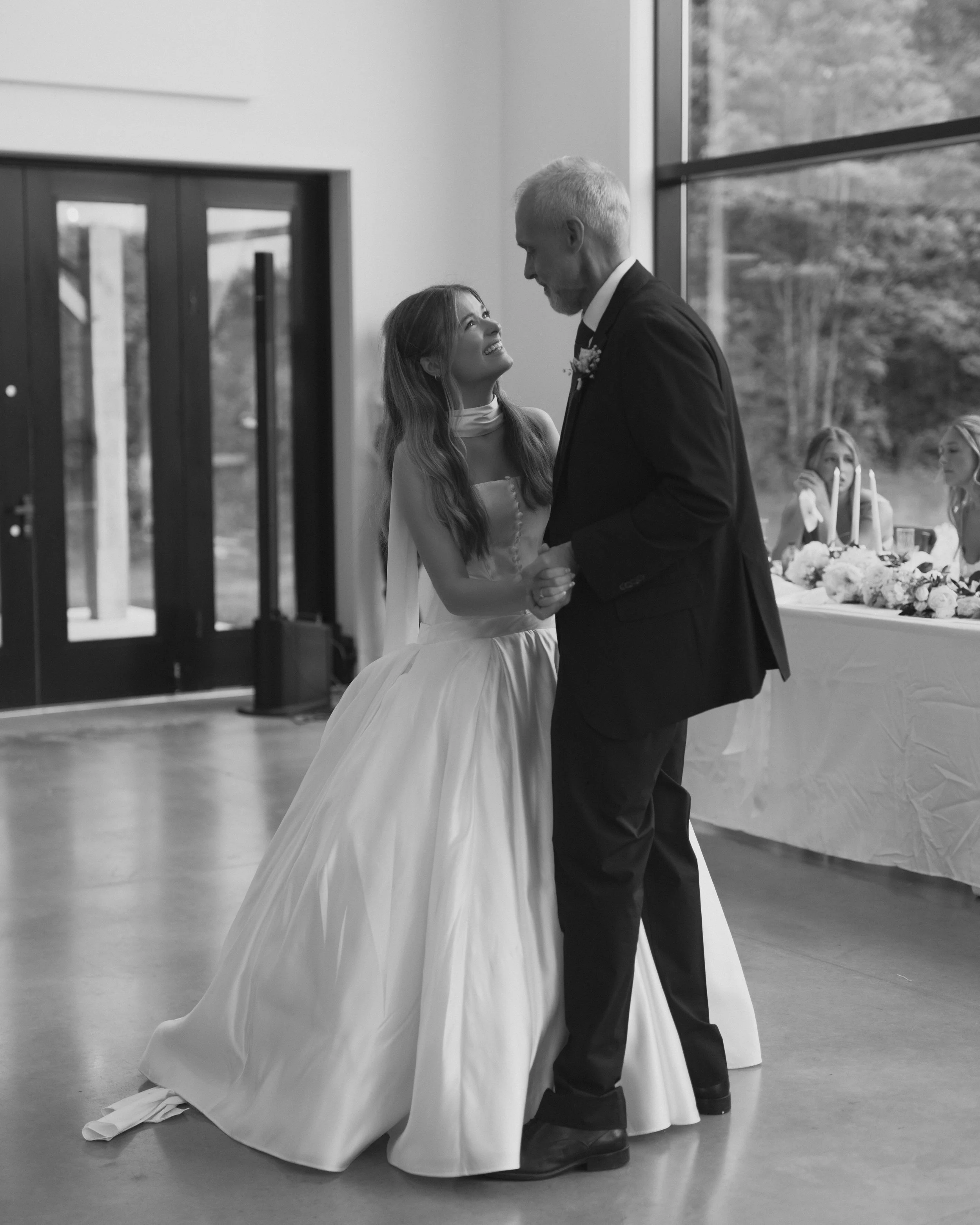 A bride and an older man, likely her father, dancing at a wedding reception. The bride is smiling up at him while they hold hands, with a decorated table and two women sitting behind them.