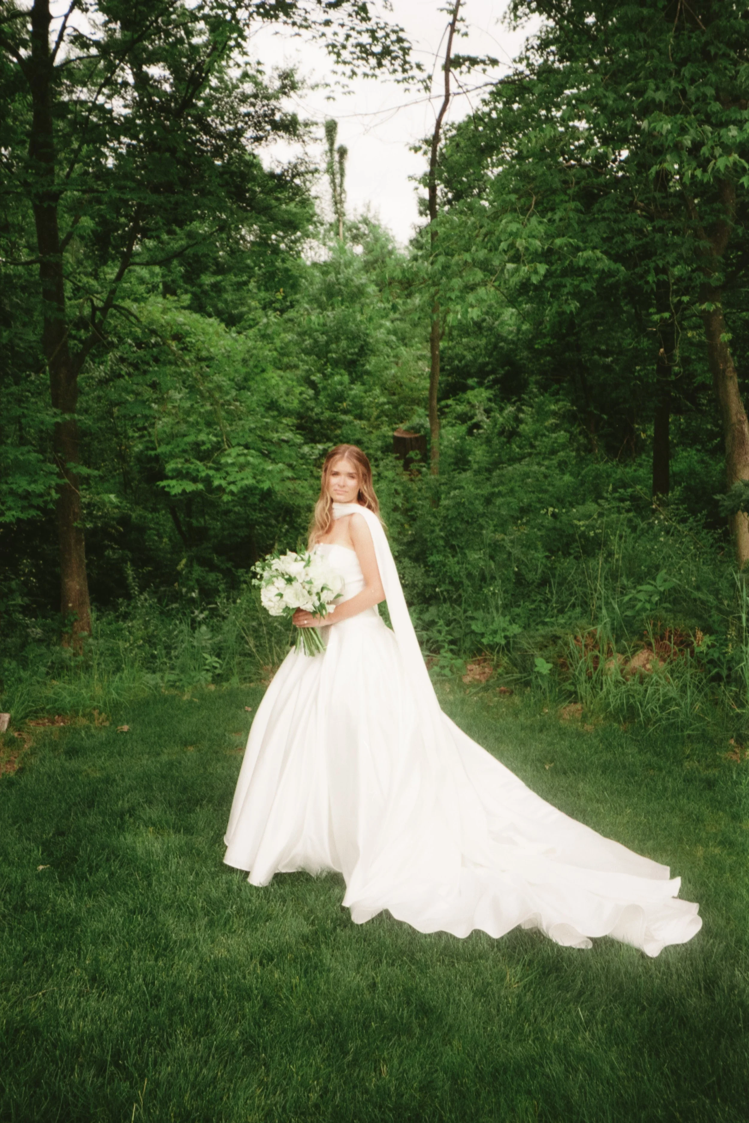 A bride in a white wedding gown holding a bouquet of white flowers, standing on a grassy area surrounded by green trees.