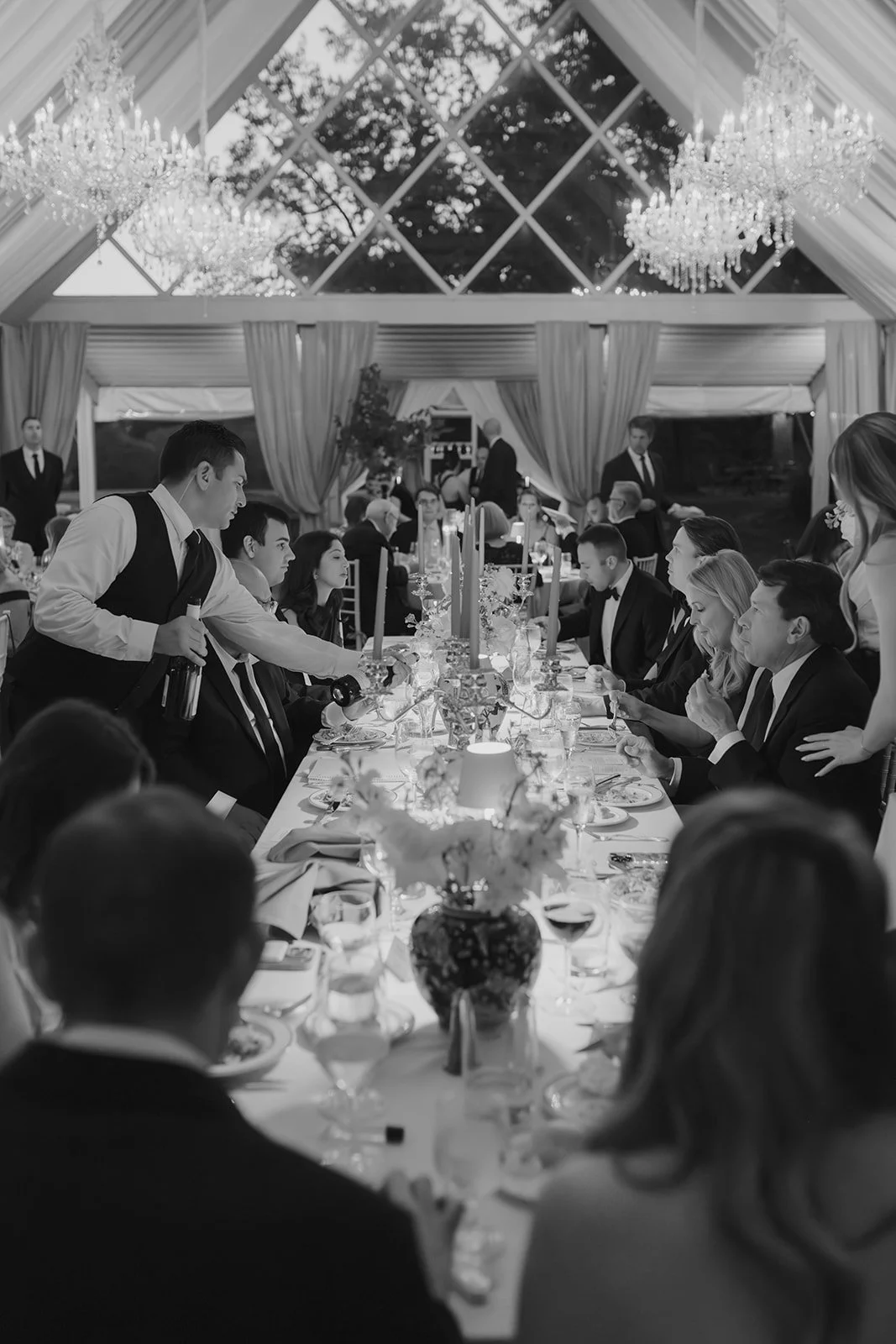 People dining at a formal banquet in an elegant tented venue with chandeliers and draped curtains, some waitstaff serving food and drink.