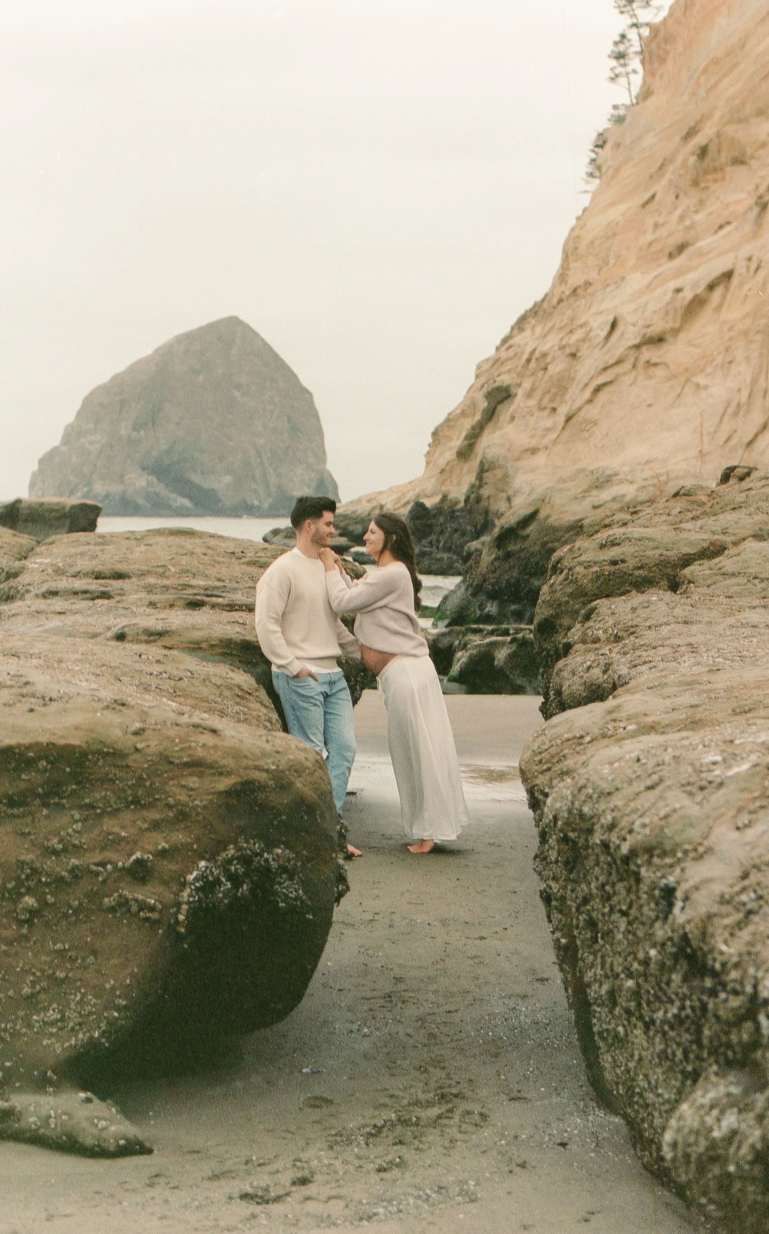 A couple standing between rocks on a beach, with a large rock formation in the background and an overcast sky.