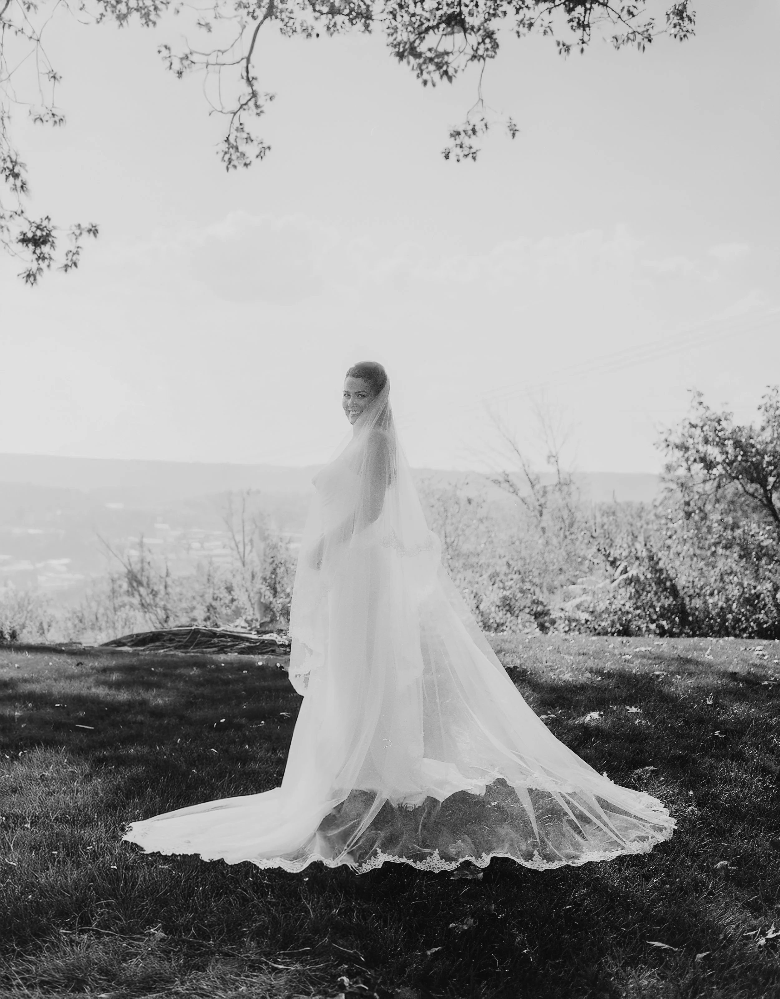 A bride in a wedding dress and veil standing outdoors on a grassy hill, smiling, with trees and a distant landscape in the background.