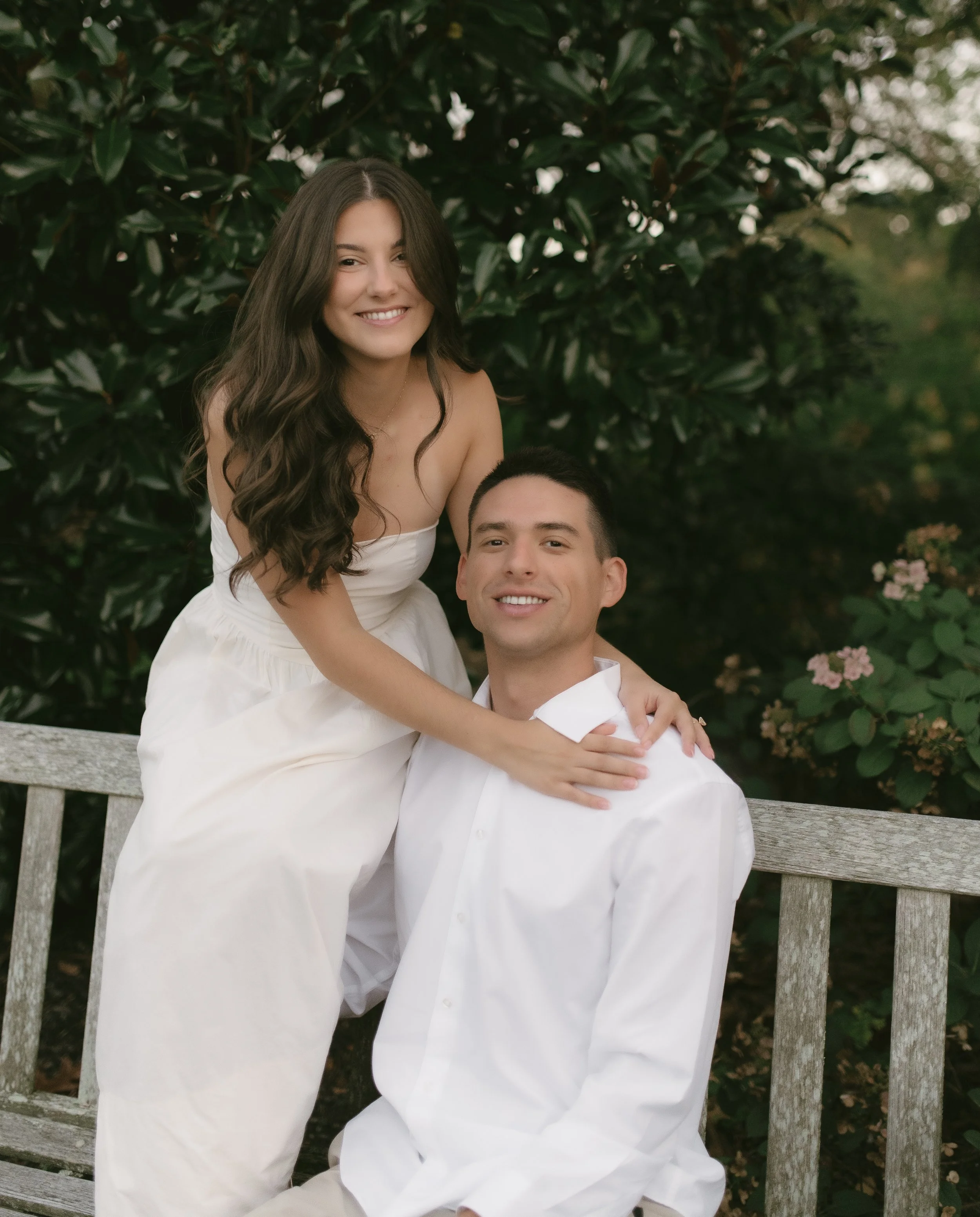 A smiling couple sitting on a wooden bench outdoors with greenery and pink flowers in the background. The woman is standing and leaning on the man's shoulder, wearing a white strapless dress. The man is sitting wearing a white shirt.