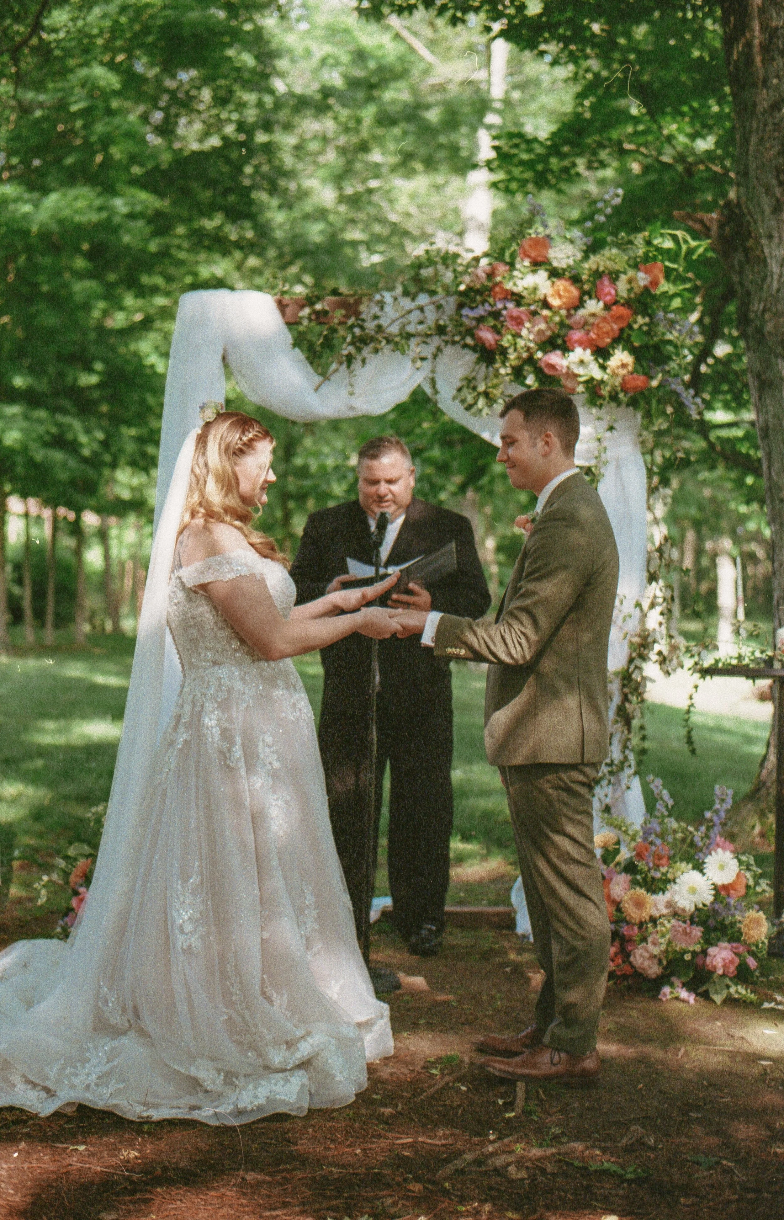 A wedding ceremony taking place outdoors in a lush green park with two people, a bride in a white wedding dress with lace details and a groom in a brown suit, exchanging vows under a decorated arch with white fabric and colorful flowers, with an offi