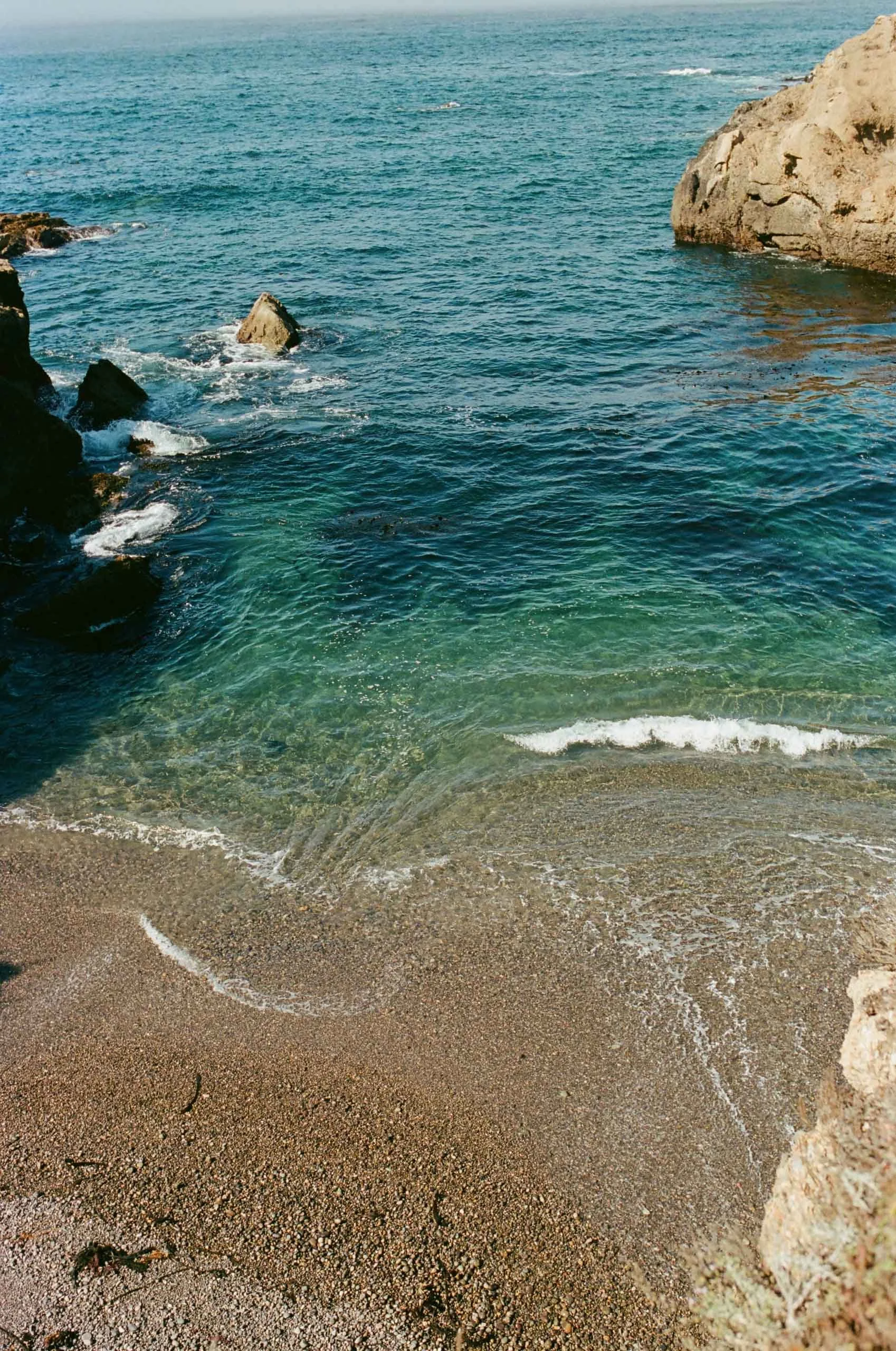 View of a sandy beach with clear blue-green ocean water gently washing onto the shore, bordered by rocky cliffs on either side.