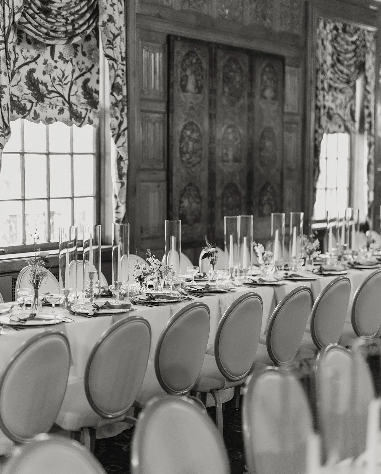 Elegant banquet table set with plates, glasses, silverware, and floral centerpieces in a historic decorated room with large windows and curtains.