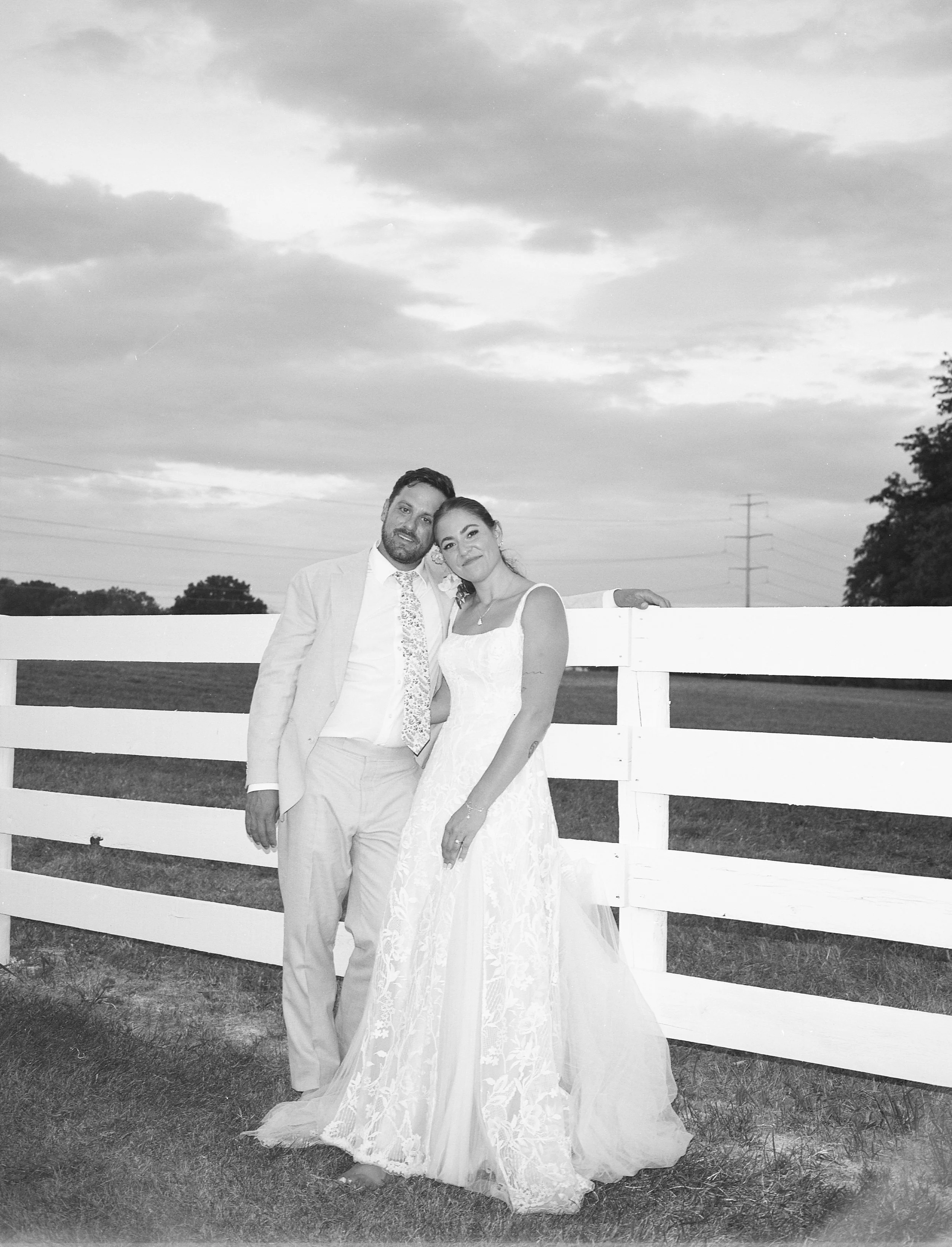A black-and-white photo of a couple dressed in wedding attire, standing close together in front of a white fence outdoors, with a cloudy sky and open field in the background.