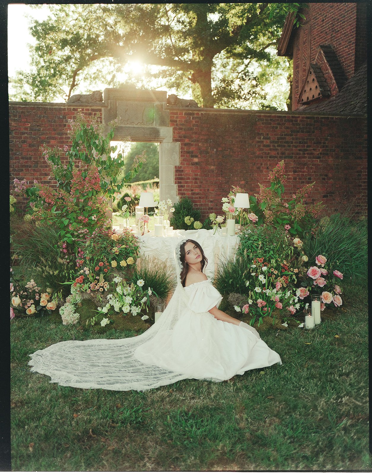 A woman dressed in a vintage white wedding gown sitting on grass in front of a floral arrangement and an outdoor wedding altar with candles, lamps, and flowers, set against a brick wall and trees with sunlight filtering through.