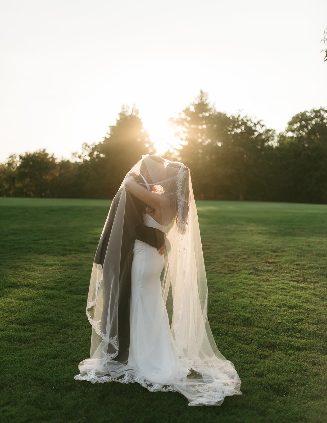 A bride and groom sharing a kiss outdoors during sunset, with the bride wearing a white wedding dress and veil, and the groom in a black suit, standing on green grass with trees in the background.