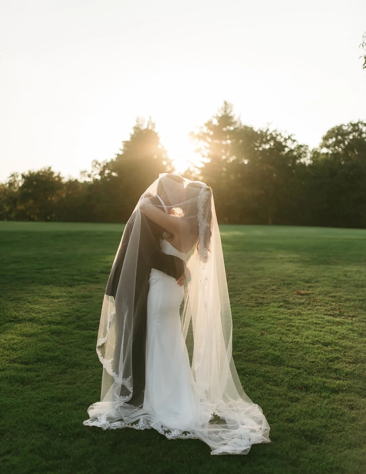 A bride and groom sharing a kiss behind a lace veil during sunset on a grassy field.