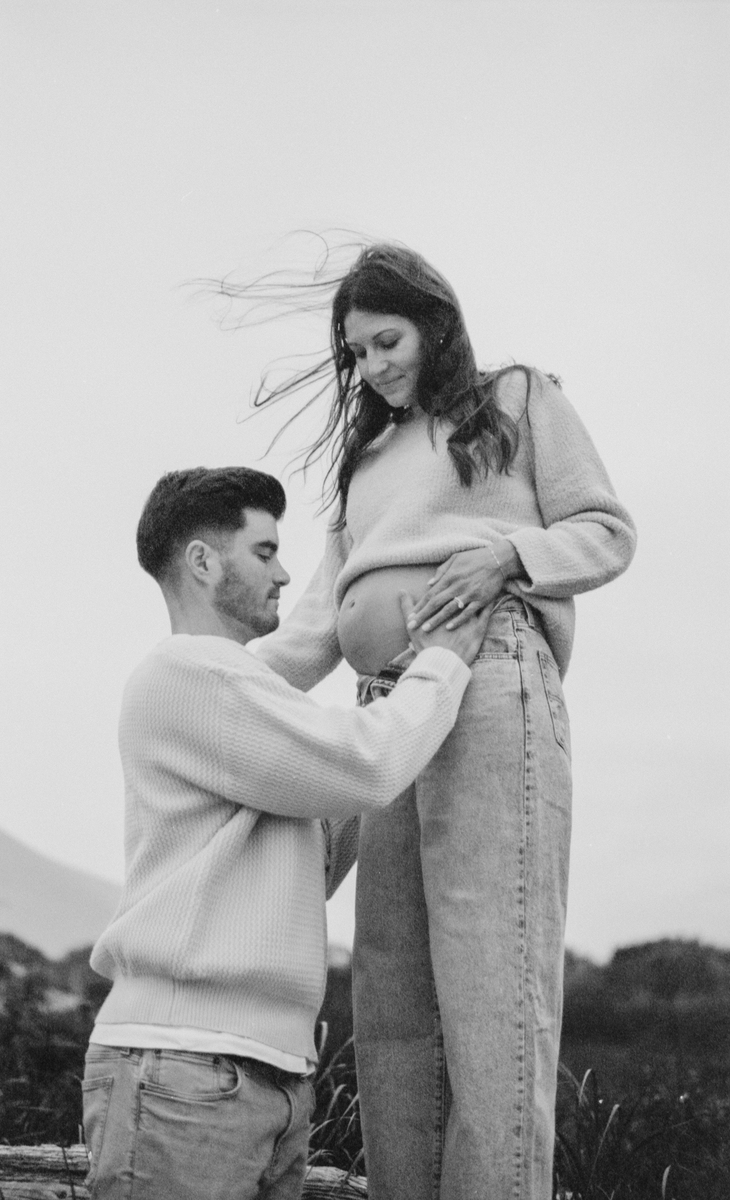 A man and a pregnant woman outdoors, with the man kneeling and touching her belly, looking peaceful. The woman has her hand on her belly and is looking down, smiling, with wind blowing her hair.