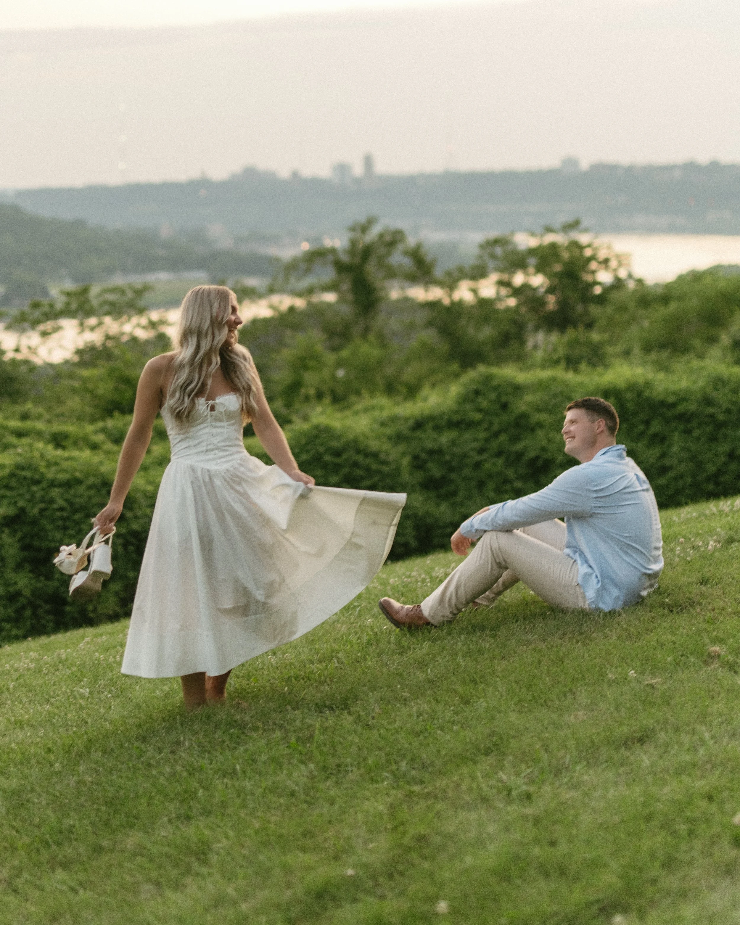 A woman in a white dress holding her shoes stands on a grassy hill, looking at a seated man in a light blue shirt who is smiling, with a green landscape and water in the background.