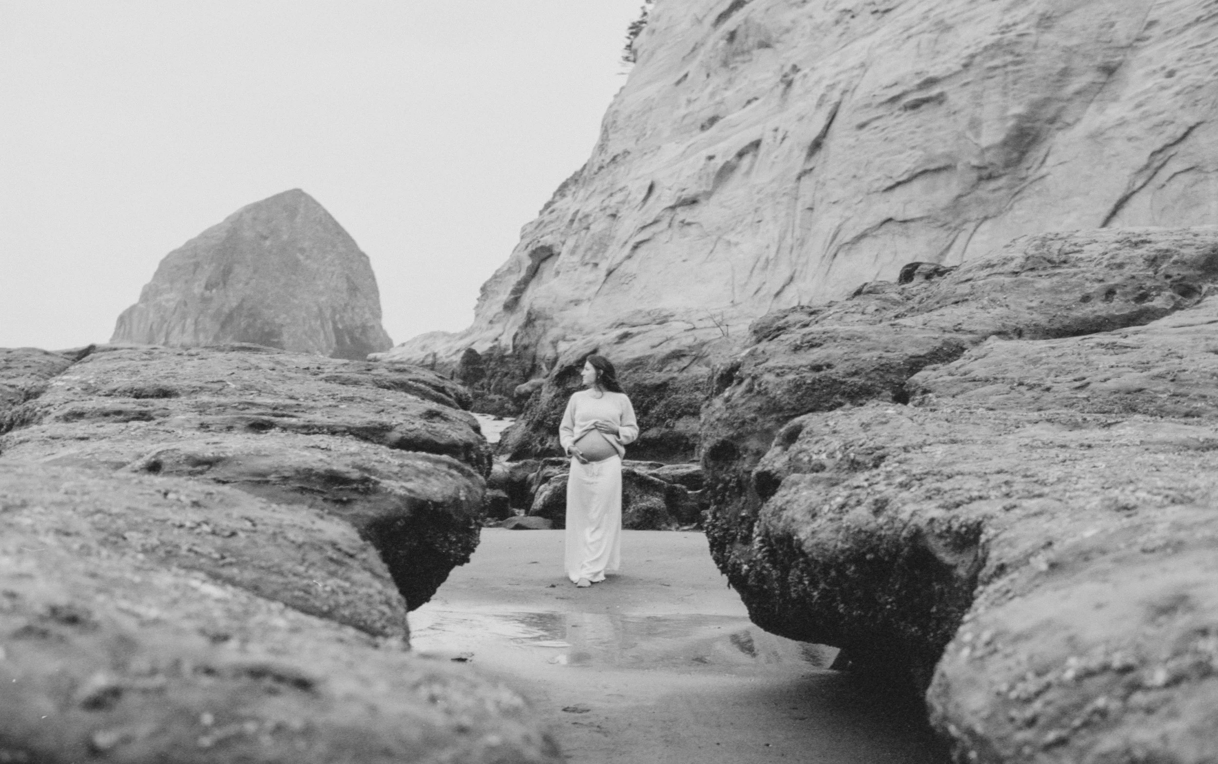 A pregnant woman standing between large rocks on a beach, with cliffs in the background, in black and white.