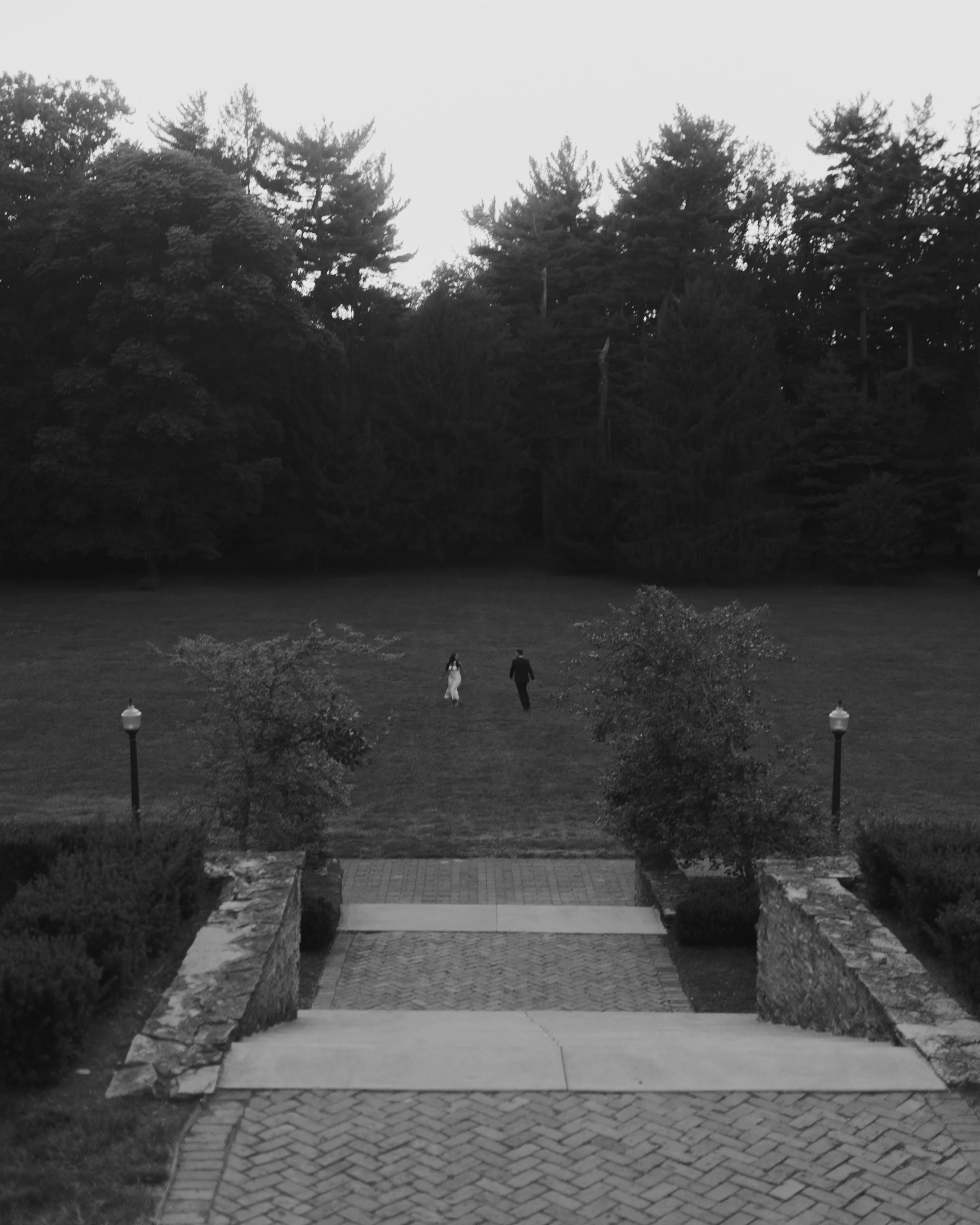 A black and white photo of a couple walking across an open grassy area towards a large wooded area in the distance, with lamps along a stone wall pathway in the foreground.