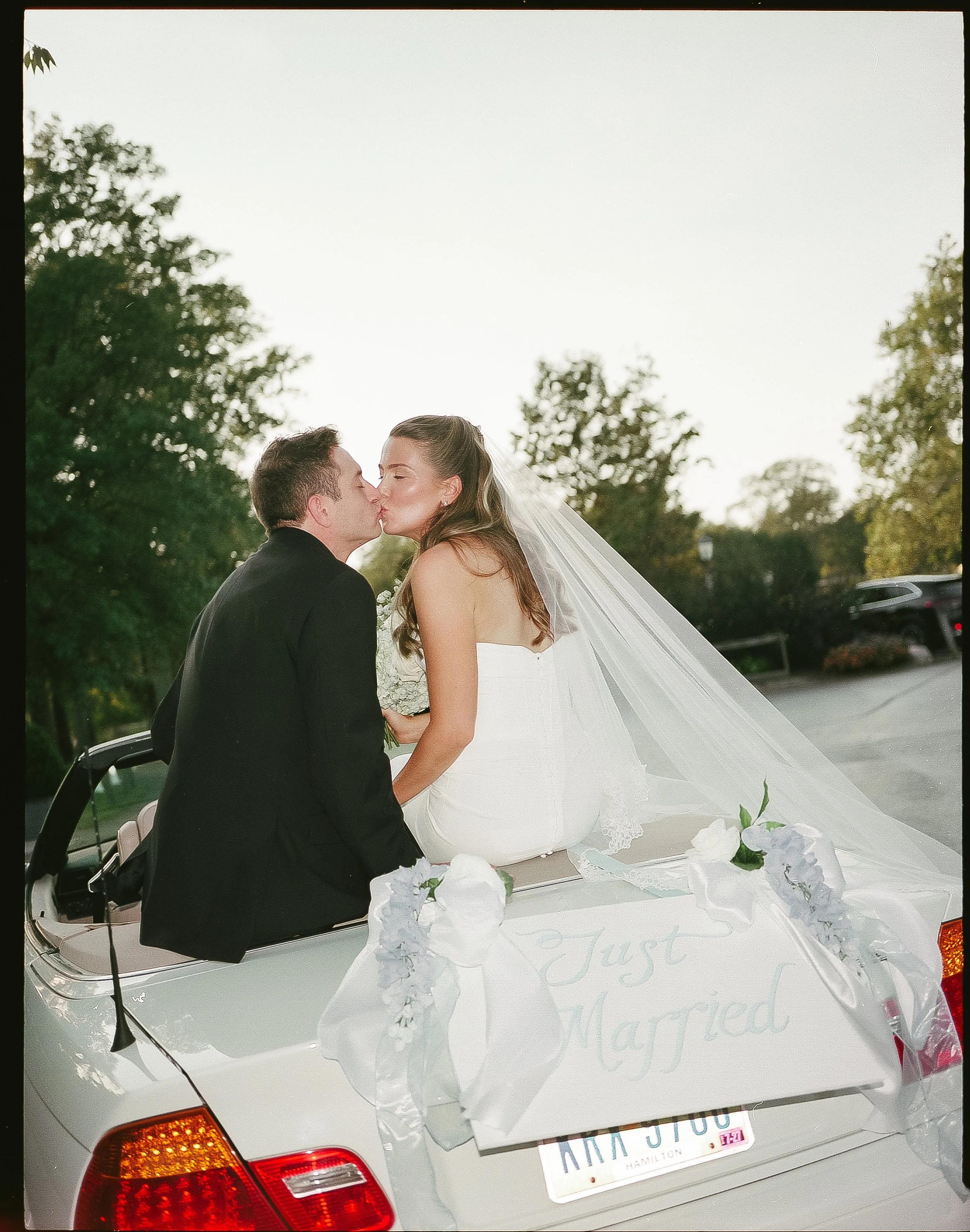 A bride and groom sitting on the back of a white convertible car decorated with white ribbons and flowers, sharing a kiss after their wedding. The car has a sign that reads 'Just Married' and a Texas license plate.