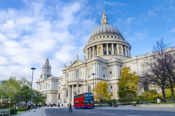 St. Paul's Cathedral in London with a red double-decker bus and pedestrians in the foreground under a blue sky.