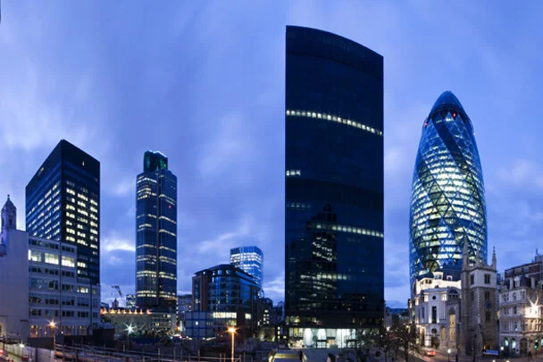 Skyline of modern skyscrapers in London, including the Gherkin building, under a partly cloudy evening sky.