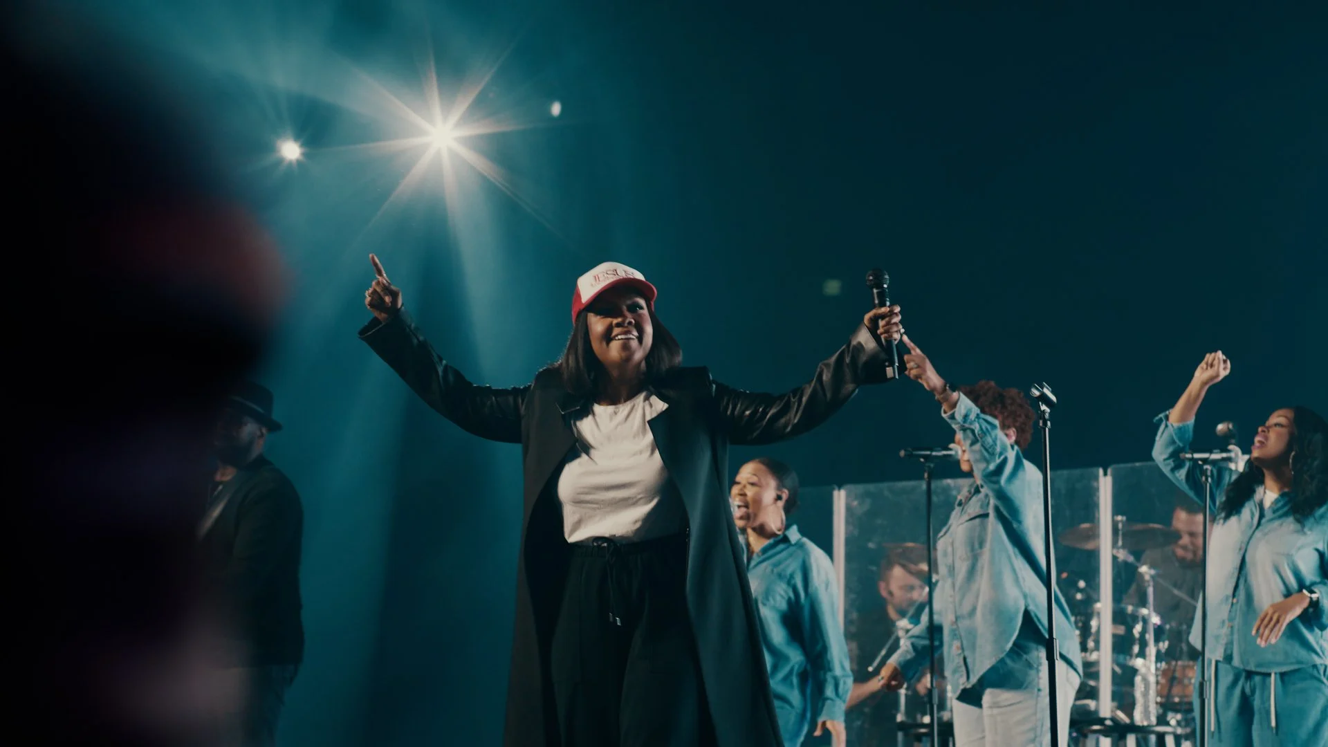 A male musician singing into a microphone on stage, wearing a black t-shirt, a black visor, and in-ear monitors, with stage lighting and a blurred colorful background.
