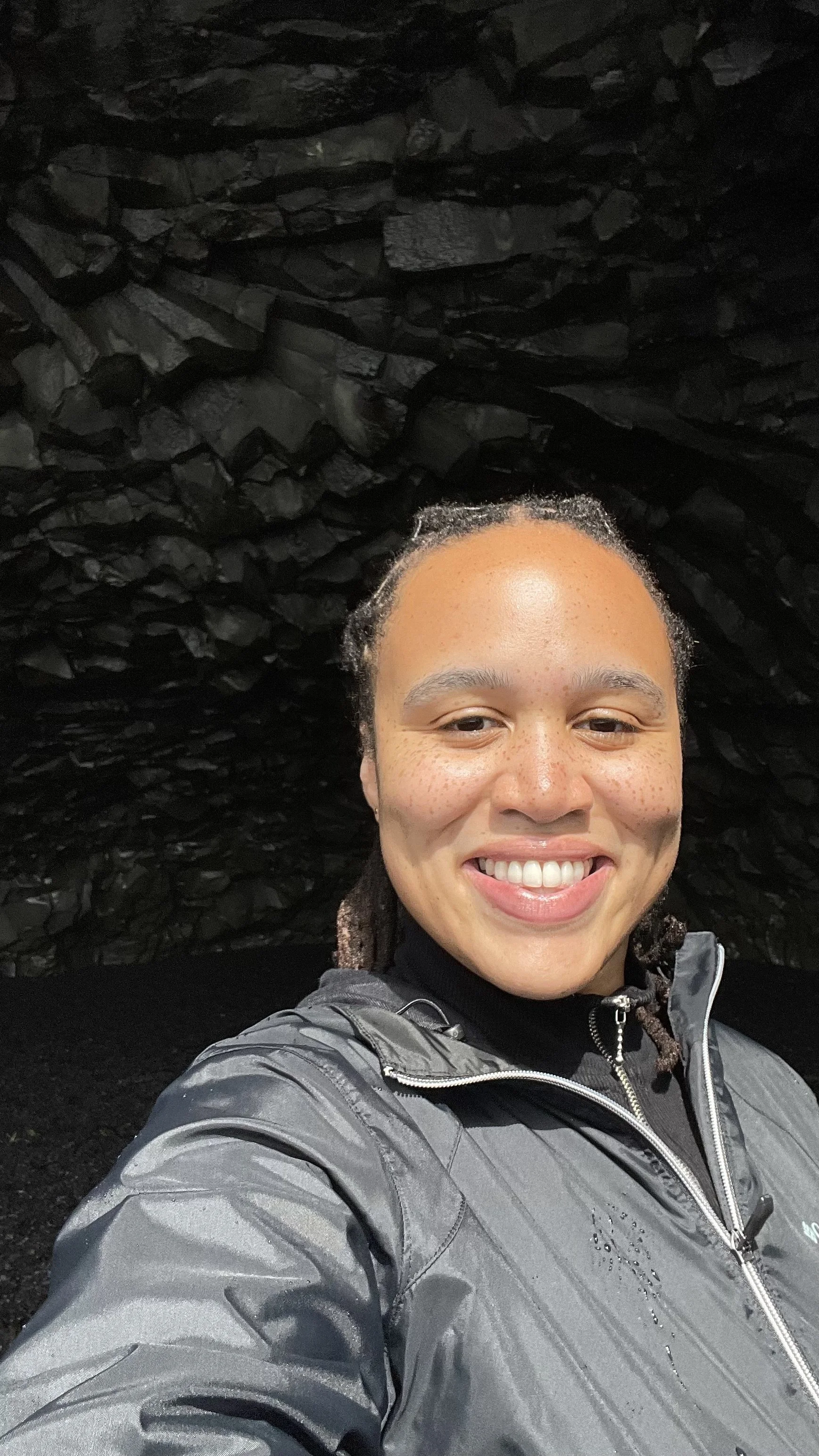 a non-binary lighter skin person smiling with locs braided straight back wearing a black zip up jacket