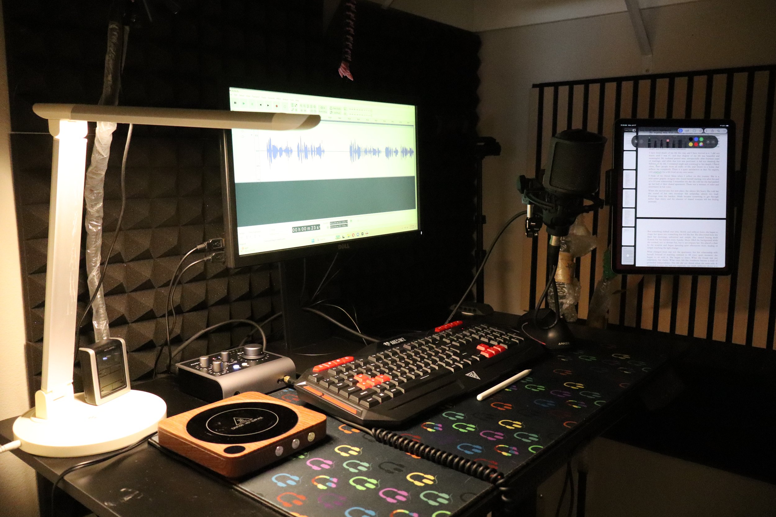 Broadcast quality home booth with sound treatment. One monitor has audio file on the screen, an IPad with script is on the side. Impeccable cable management with the microphone, keyboard, audient i4 interface and most important tea mug warmer.