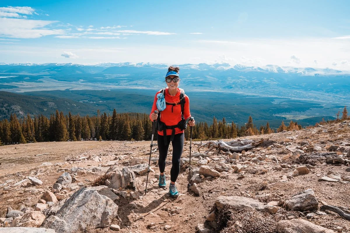 a woman hiking up mount elbert in colorado with a bright sun hoodie and trekking poles