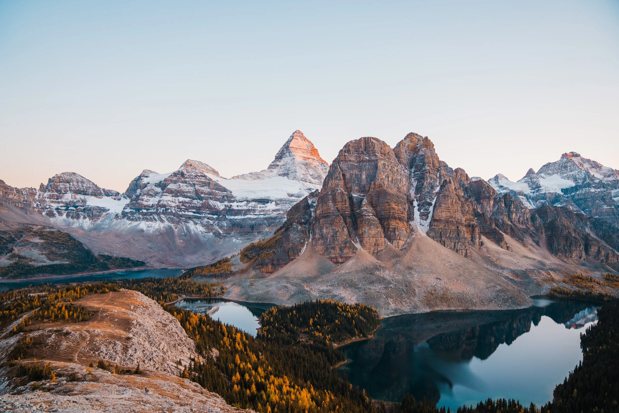 Snow-capped mountains with rugged peaks overlooking two calm, reflective lakes, surrounded by forested slopes and a clear sky.
