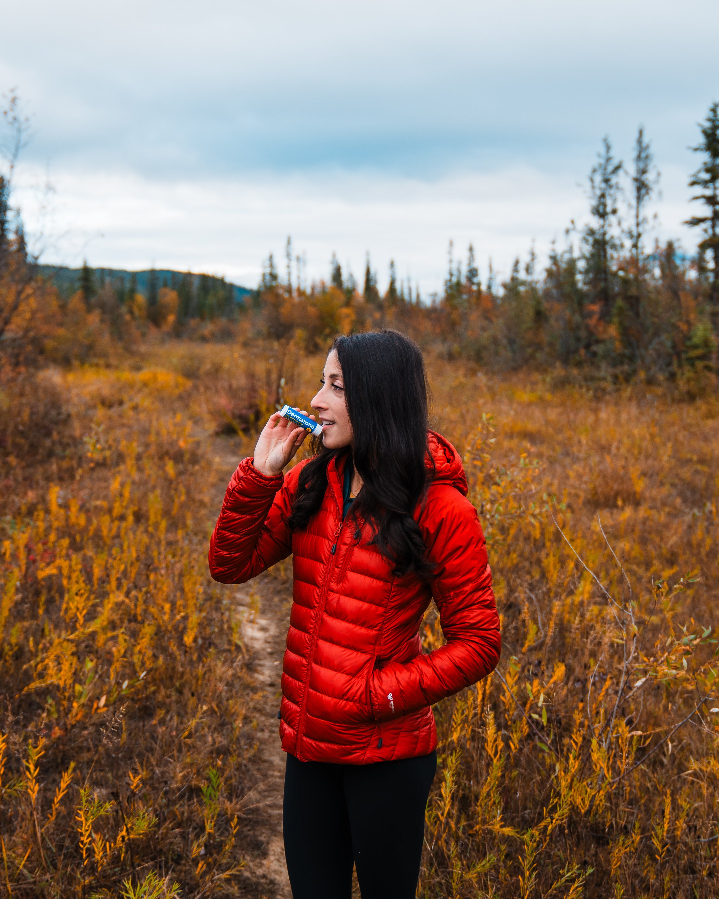 A woman in a red puffer jacket holding a lip balm in a natural outdoor setting with autumn foliage.