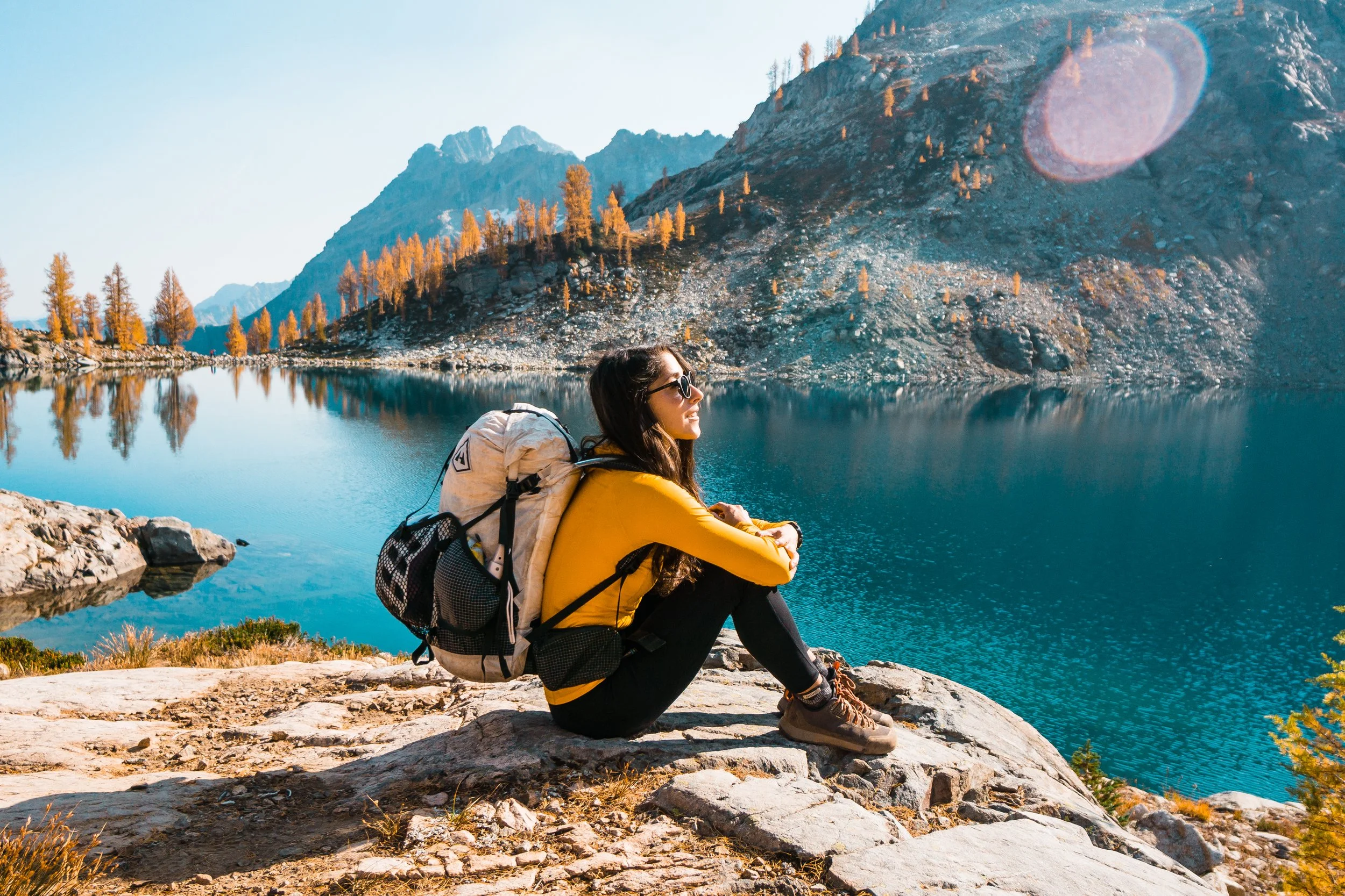 A woman sitting on a rock by a mountain lake with trees and mountains in the background, wearing a yellow jacket, hiking boots, sunglasses, and carrying a backpack.