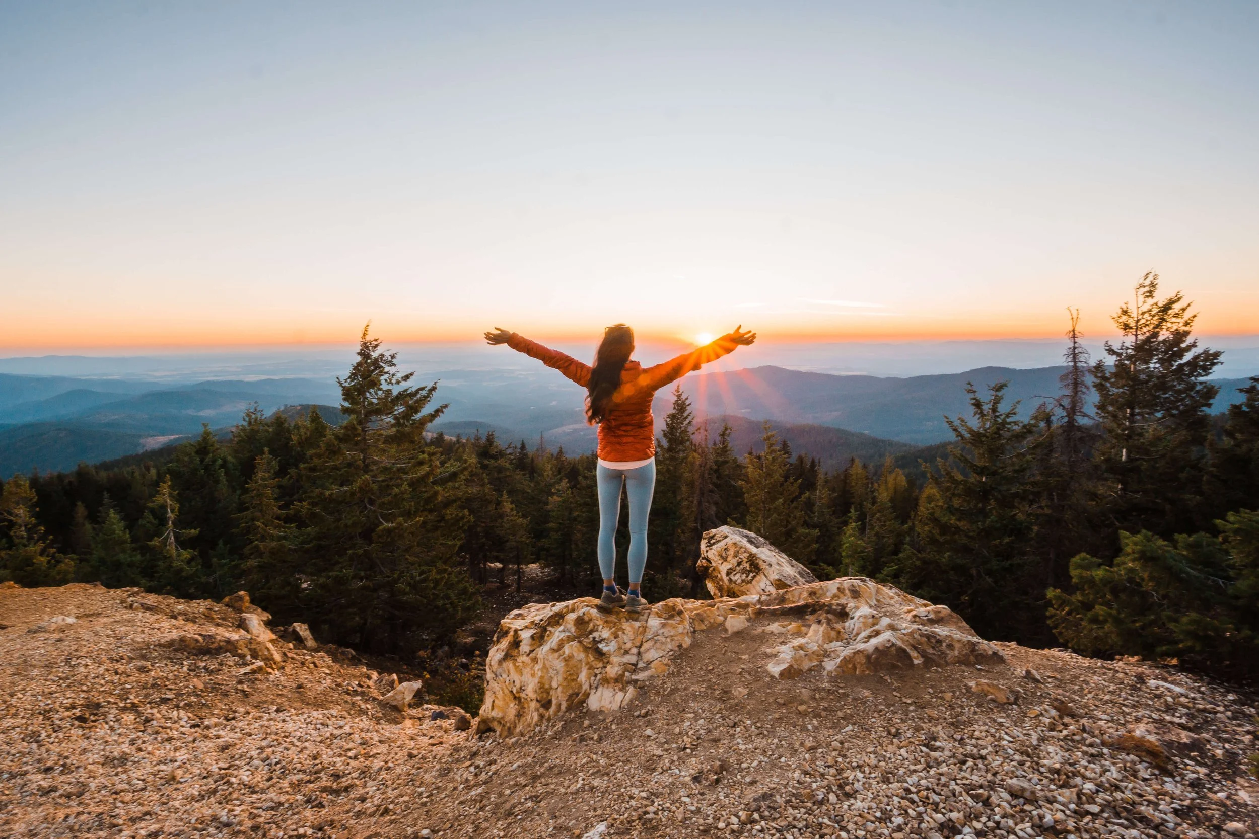 A woman standing on a rocky mountain ledge with arms raised, watching the sunset over a forest of evergreen trees and distant mountains.