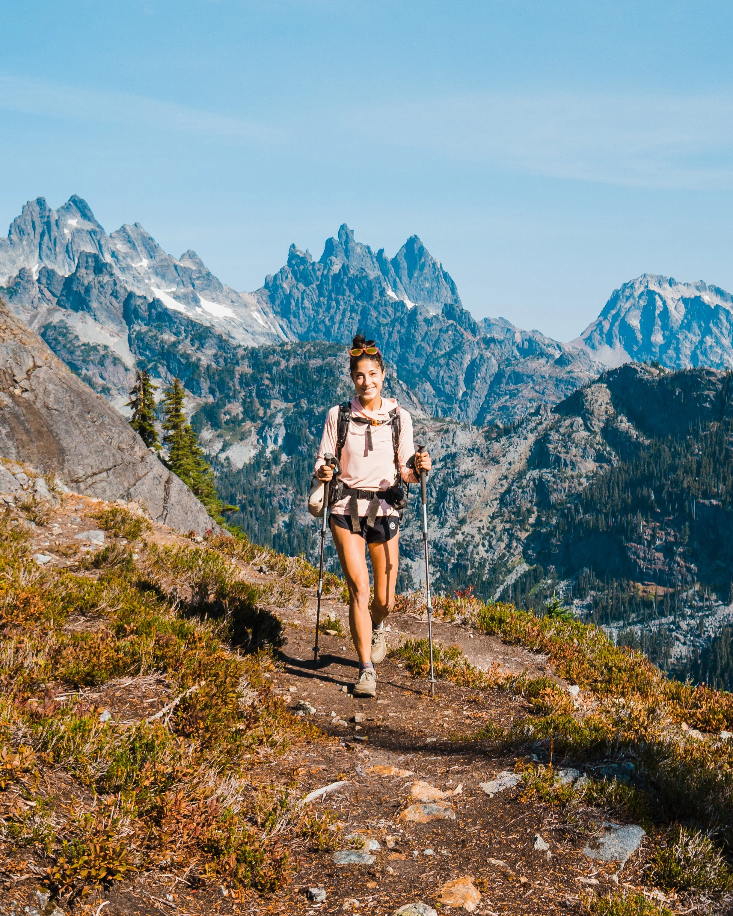 A woman hiking on a mountain trail with a scenic view of rugged peaks and blue sky in the background.