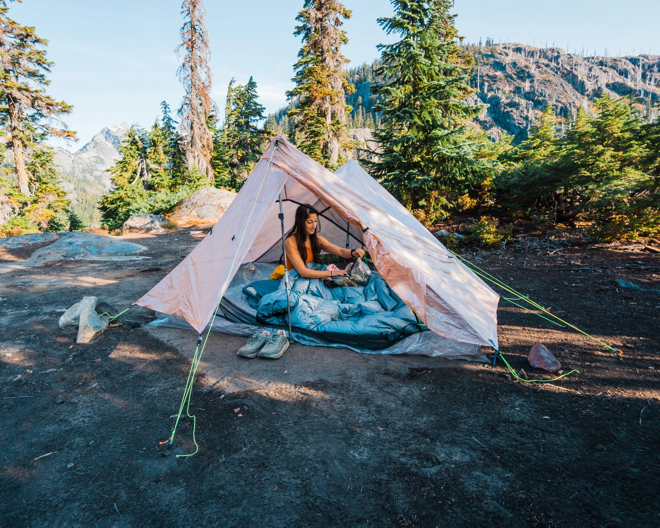 A woman inside a camping tent set up on a forested mountain area with tall trees, rocky terrain, and mountains in the background, preparing her gear.
