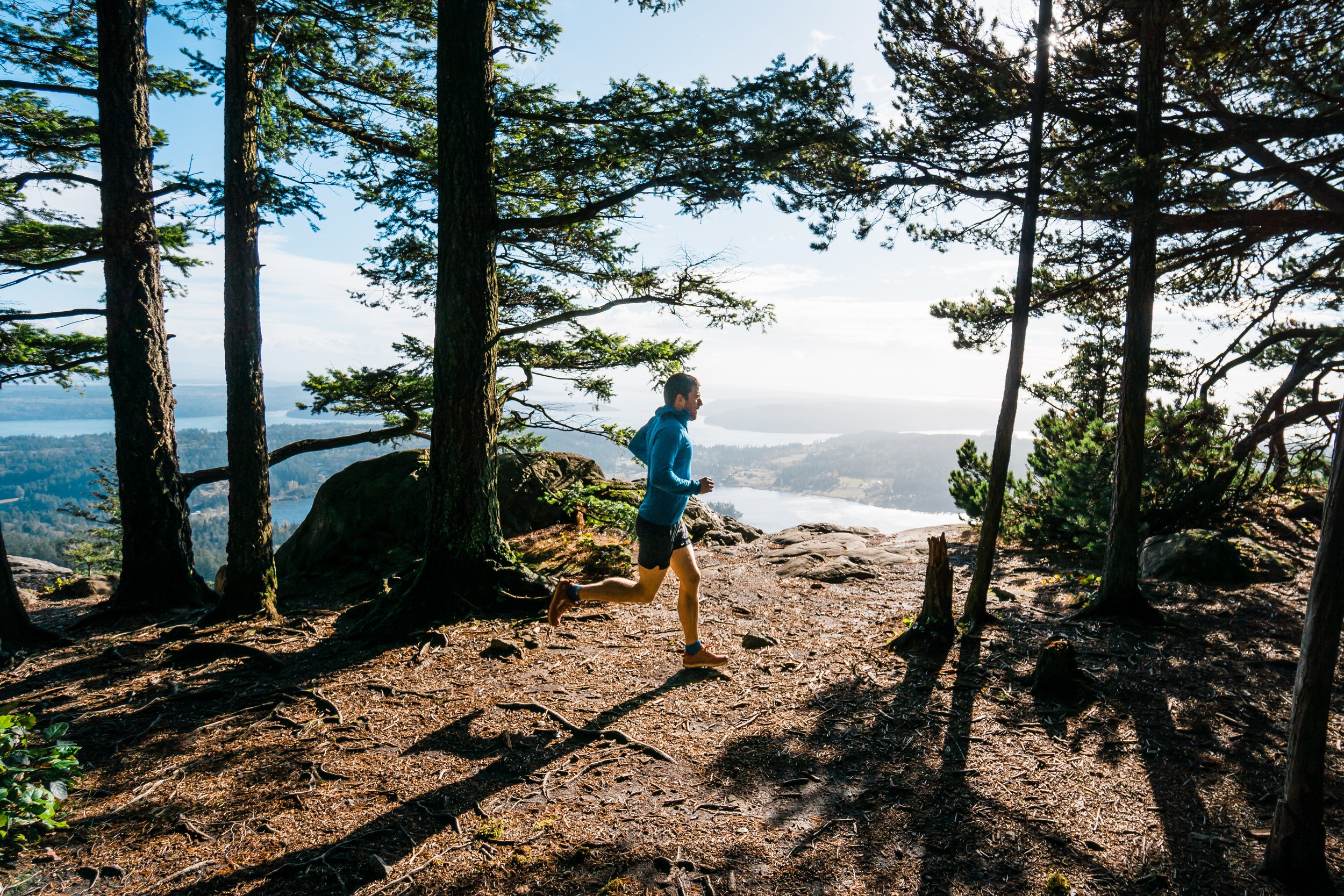 A person running on a trail surrounded by trees with a scenic view of water and mountains in the background.
