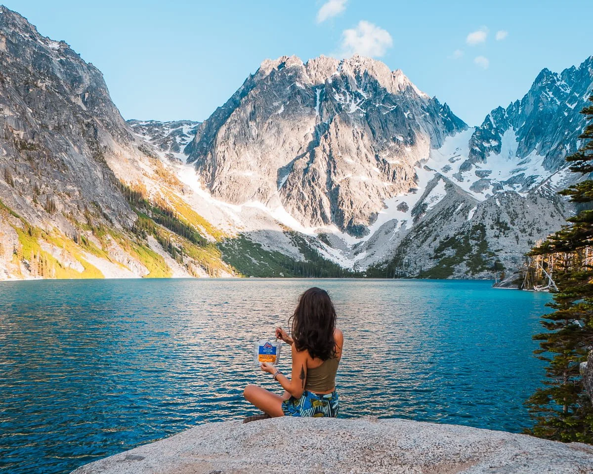 a woman sitting on a rock in front of an alpine lake, eating a freeze dried meal with jagged peaks in the background