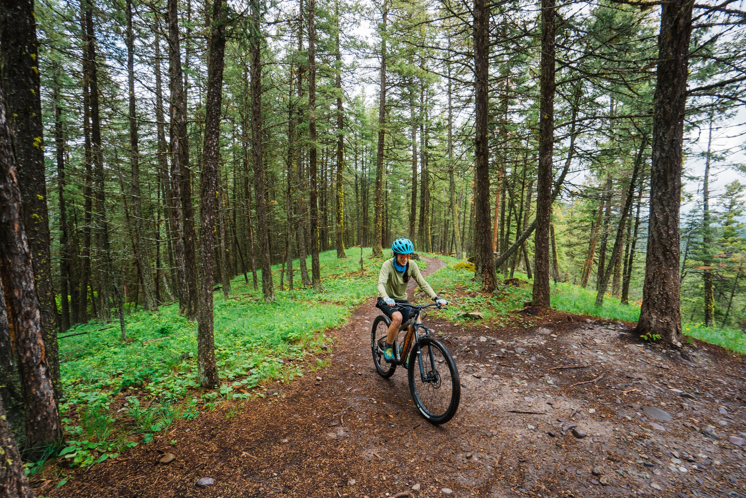 Person riding a mountain bike on a dirt trail through a green forest with tall trees and a cloudy sky.