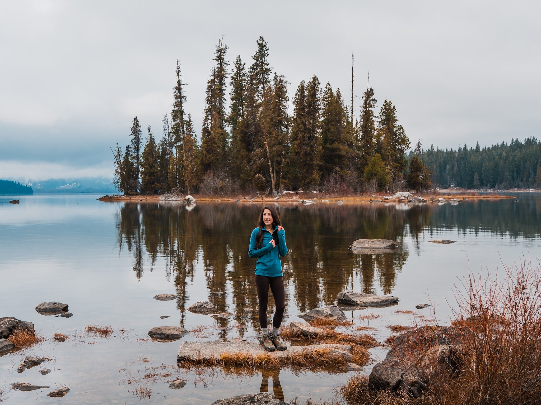 a woman wearing a blue fleece and dark leggings, standing on a rock in front of a lake with a large, forested island in the background