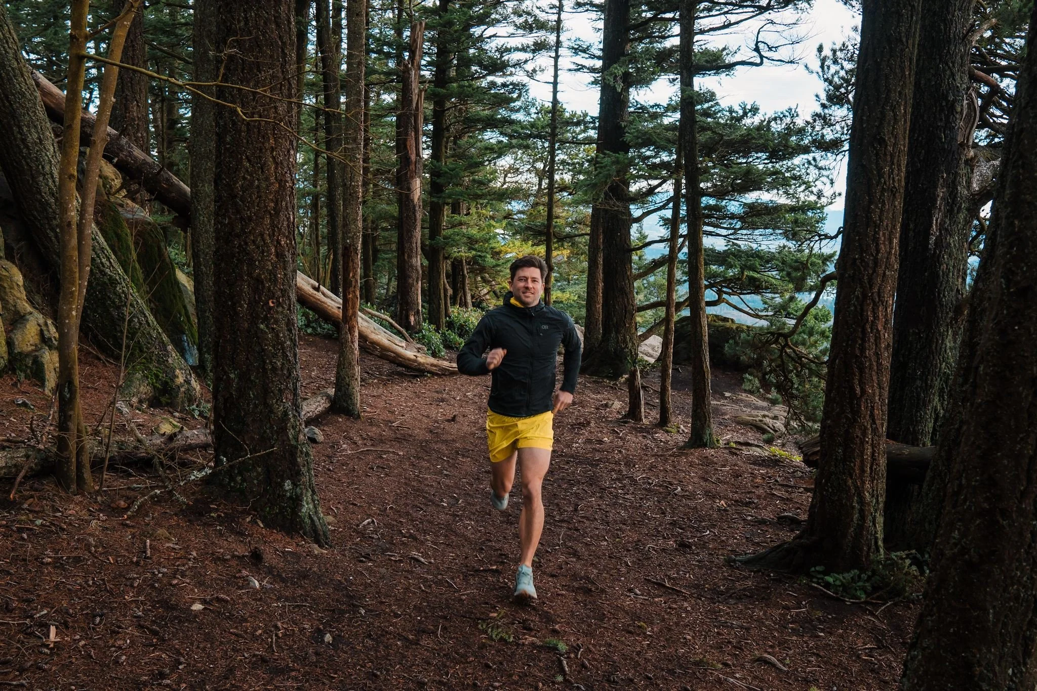 A man trail running through a forest, wearing a black jacket and neon yellow shorts