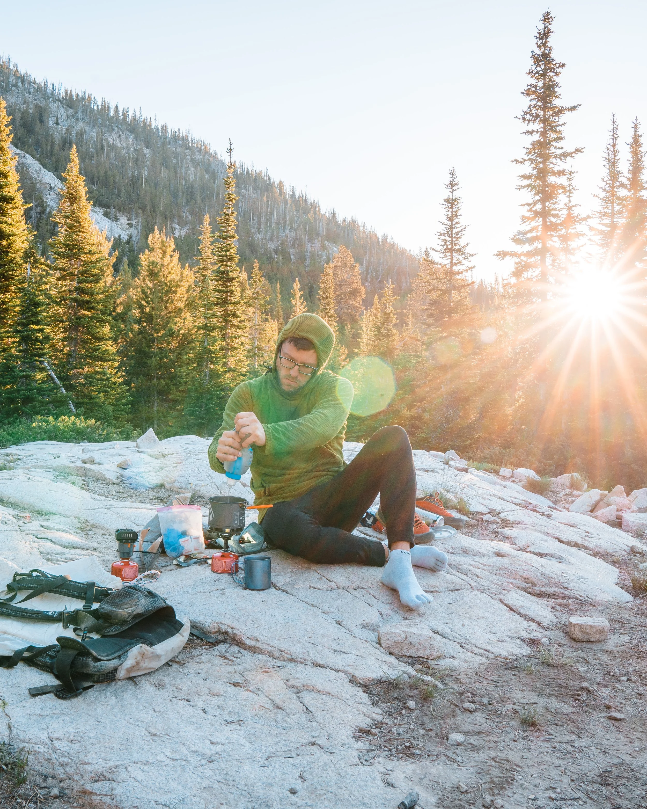 A person in a green hoodie and glasses camping outdoors on a rocky surface in a forest with tall pine trees, cooking with a portable stove, as the sun sets behind the trees.