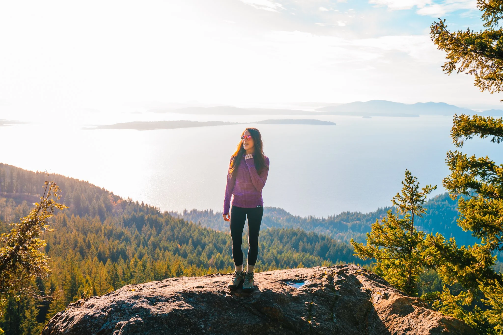 a women standing on a rocky outcropping, overlooking water, pine trees, and islands
