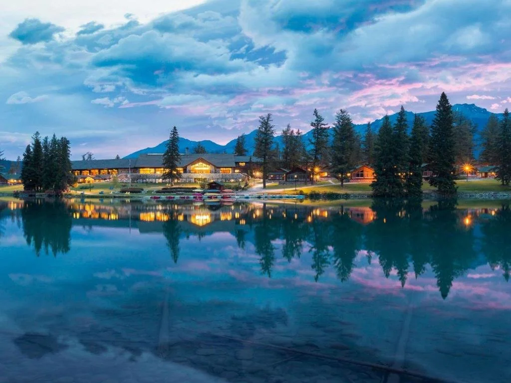 A lodge sitting on the edge of a reflective lake with trees and mountains in the background. This is the Fairmont Jasper Park Lodge