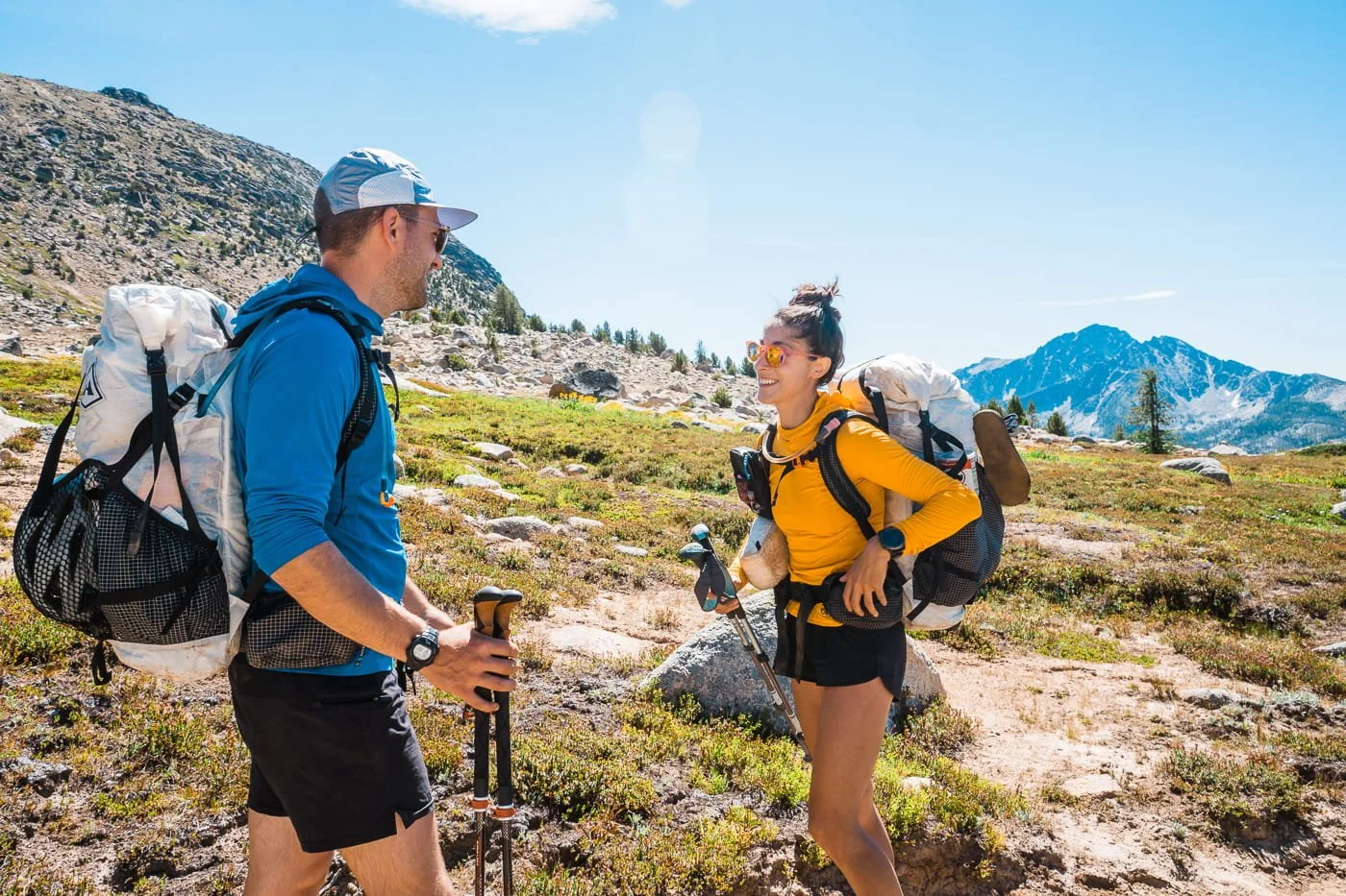 a man and woman backpackers, both wearing bright colored sun hoodies and shorts on the trail