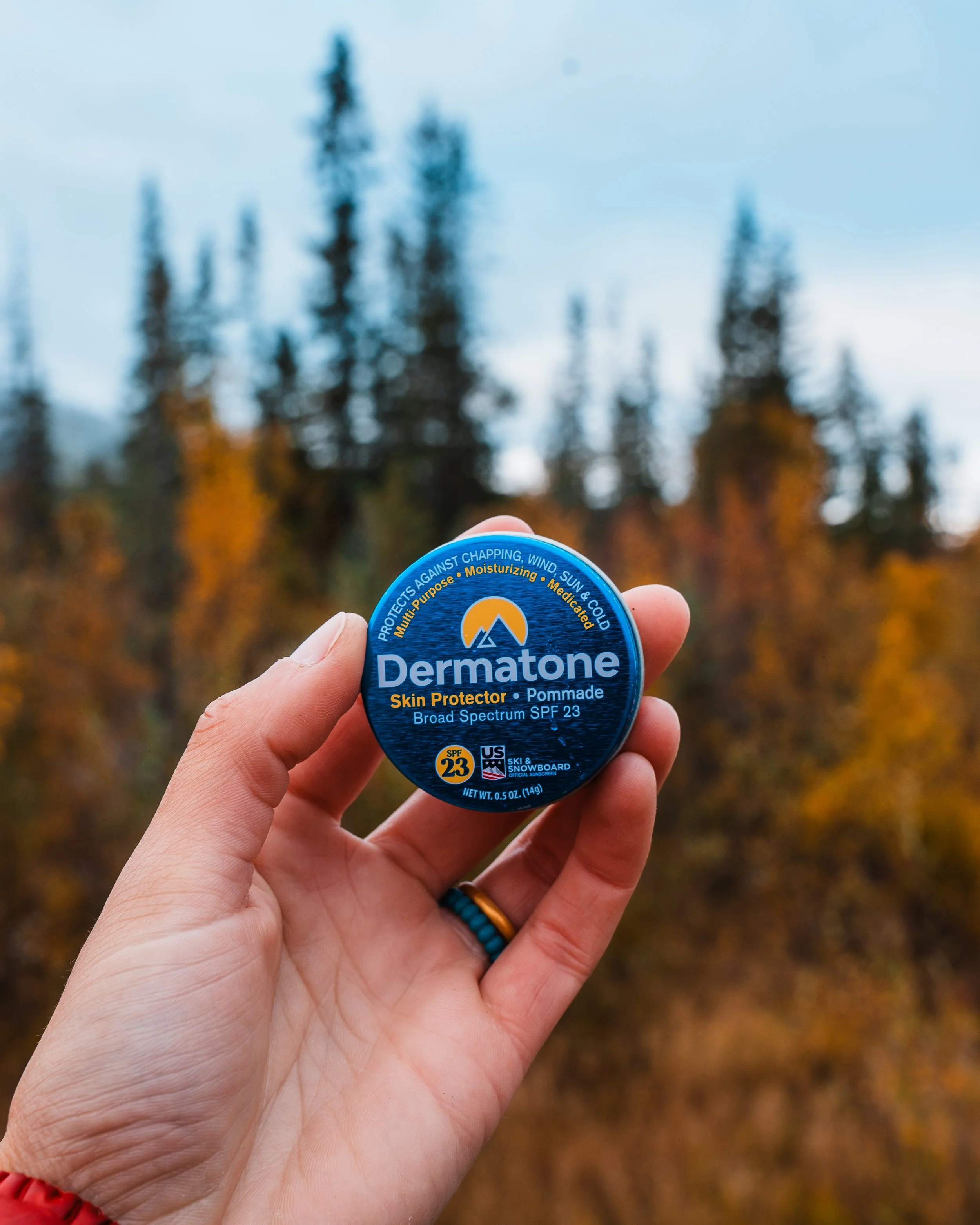Hand holding a blue container of Dermatone skin protector with a forest of autumn trees in the background.